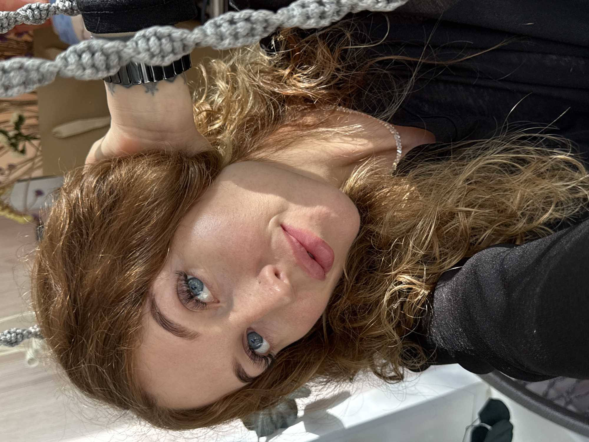 The image is a selfie taken indoors, likely in an apartment, based on the light and décor. The selfie shows a fair-skinned woman with strawberry blonde hair and blue eyes, with her head tilted and angled toward the camera. She wears black clothing. A grey, knotted rope or cord-like object is visible in the upper left corner, near a wrist sporting a black watch. A faint tattoo can be seen on the wrist. The woman has styled eyebrows and appears to be wearing light makeup including lip color and eyelash extensions. The lighting is strong and appears to be mostly from a natural source. The presence of the gray rope in the upper frame suggests she may be on or near a piece of furniture. There is insufficient data to determine the location of the city of Chaikovsky, Russia.