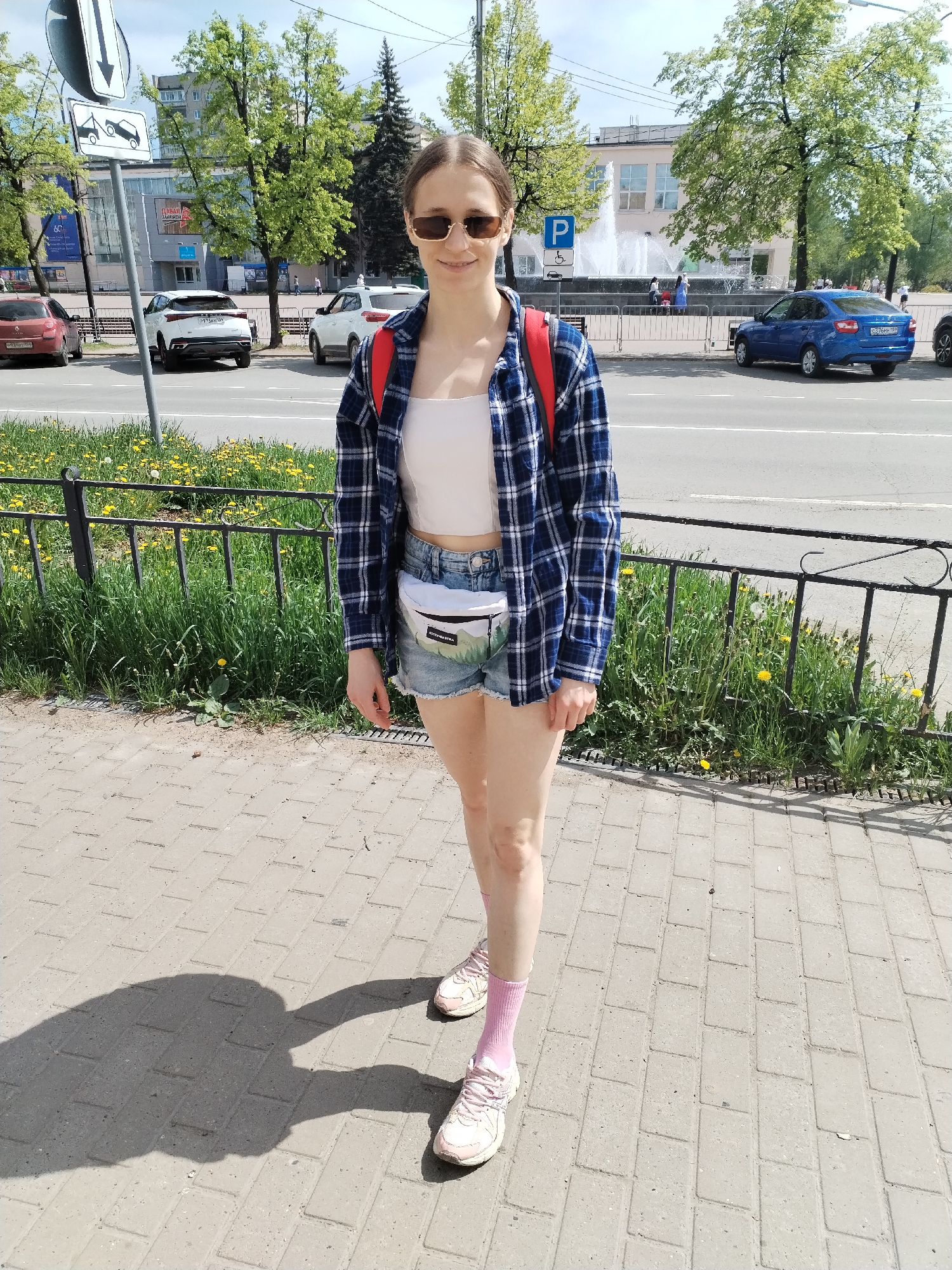 A woman stands on a brick sidewalk in Chaikovsky, Russia. She wears sunglasses, a white tank top, denim shorts, a blue plaid shirt, a white patterned waist pack, pink socks, and pink sneakers. A shadow of a person is visible on the pavement to the left. Behind her, there is a black metal fence with grass and dandelions growing on the other side. A road separates the sidewalk from a parking area with several cars, including a red hatchback, a white SUV, and a blue sedan. Across the road, there is a fountain, a parking sign, trees, and buildings. A sign with the image of a vehicle being towed is visible in the background.