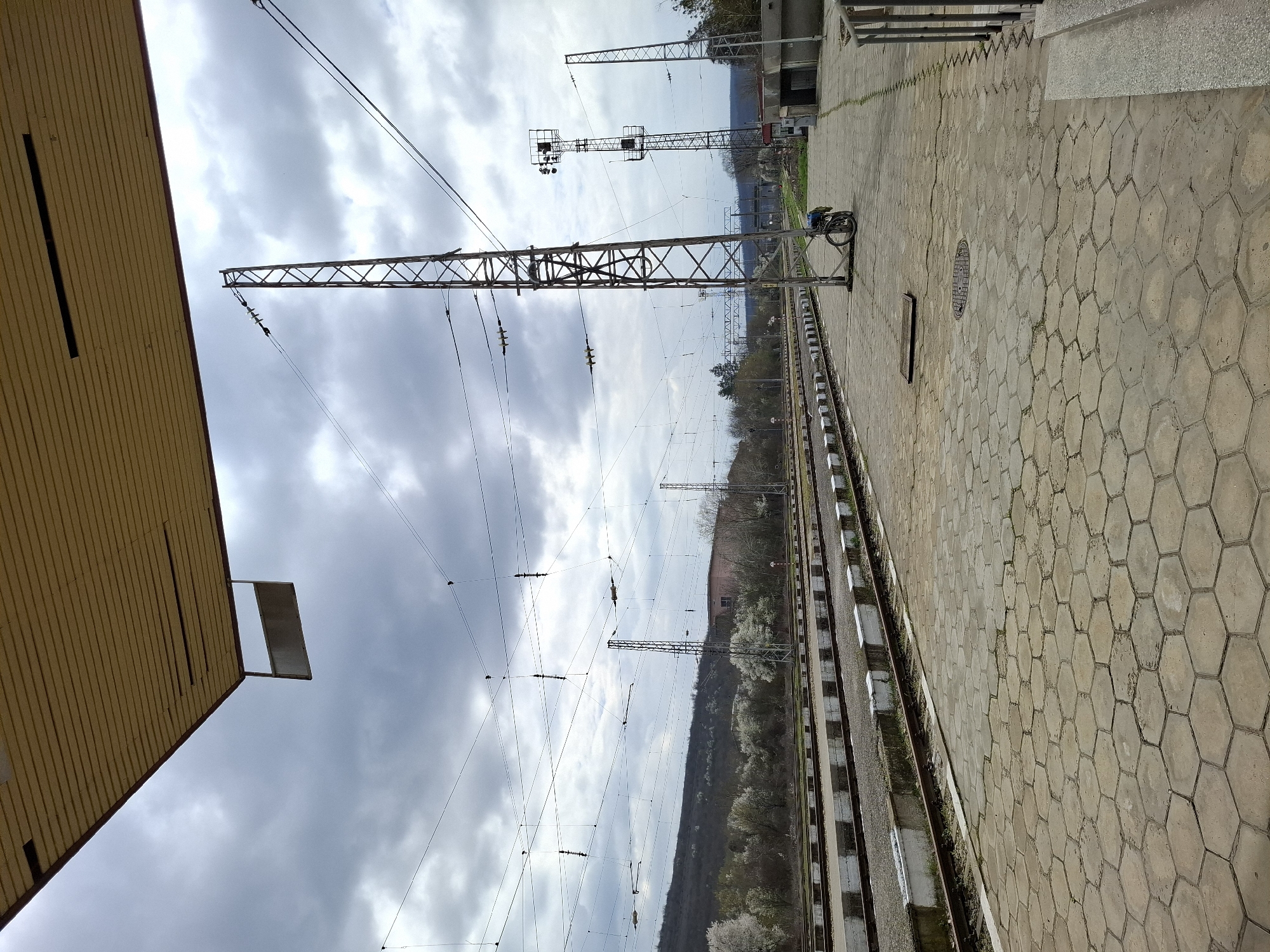 The image shows a railway station in Strazhitsa, Bulgaria. The platform is paved with hexagonal concrete blocks. Railway tracks run alongside the platform. Overhead power lines and supporting structures are visible. A station building is partially visible to the left. Trees and hills are visible in the background. A bicycle is parked on the platform. The sky is cloudy.