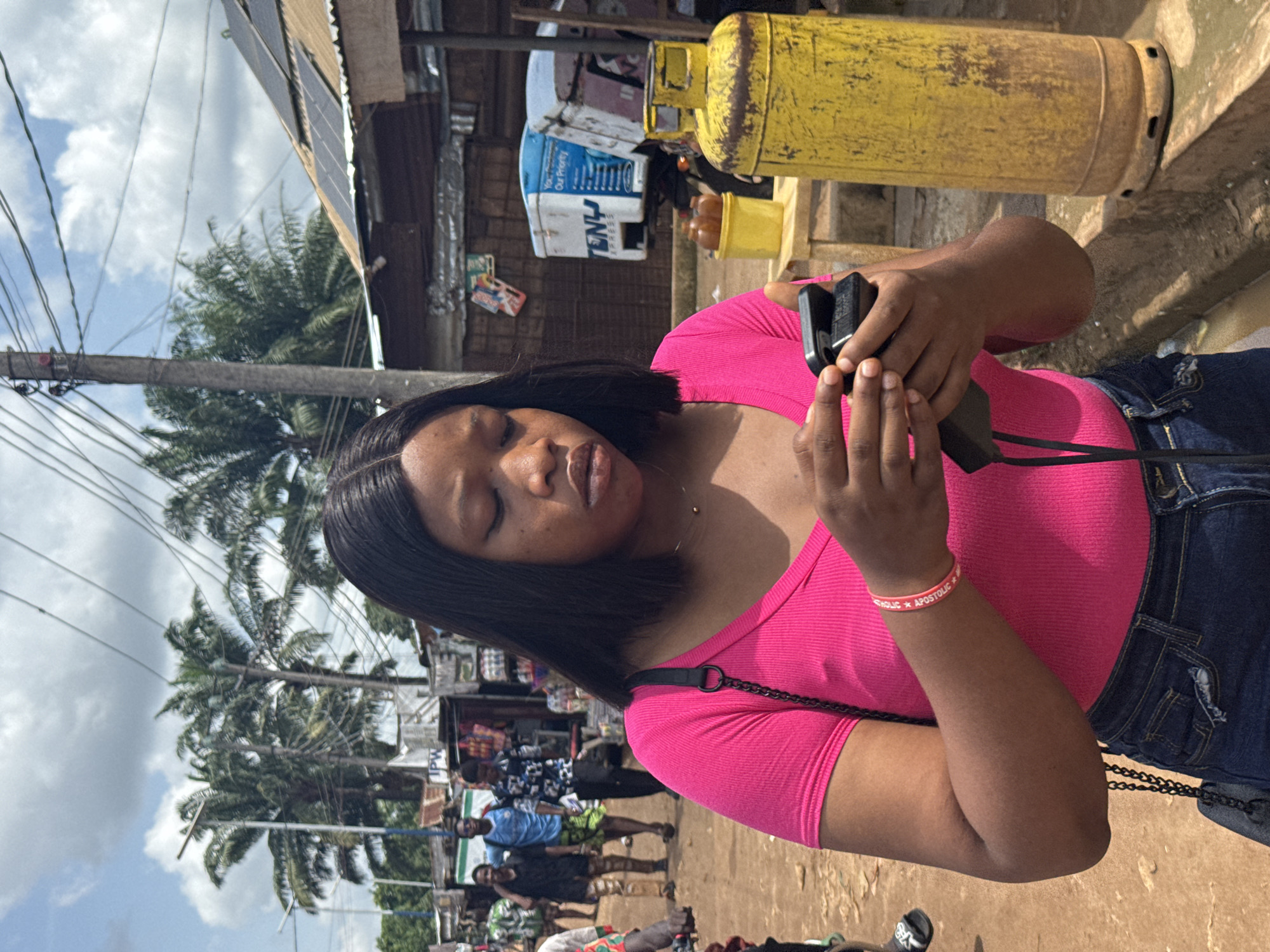 The image, taken in Ihiagwa, Nigeria, captures a vibrant street scene during daylight hours. A young woman is the central subject, positioned in the foreground and slightly to the right, with the photo appearing rotated 90 degrees clockwise from its natural orientation. She has dark, straight hair and wears a bright pink ribbed v-neck top and blue denim jeans. On her right wrist, she wears a pink wristband inscribed with the partial text "CATHOLIC * APOSTOLIC *". She is holding a black electronic device, likely a camera or smartphone, with both hands, looking down at it with a focused expression and slightly parted lips. A black strap is draped across her body.
The background depicts a bustling outdoor environment. On the left, numerous tall palm trees are visible under a partly cloudy sky, crisscrossed by overhead power lines and a utility pole. On the right, various structures with corrugated metal roofs are present, one of which features solar panels. Two large, yellow, rusted gas cylinders stand upright on a raised platform near these buildings. Below them, a blue and white vending machine or kiosk is visible, displaying the text "TENJ" or "TENU", "EXPRESS", and "Our Priority". In the deeper background, several people are seen walking along what appears to be a busy street or market area. The ground is a light brown, unpaved surface. The scene conveys the lively ambiance of a typical Nigerian street.