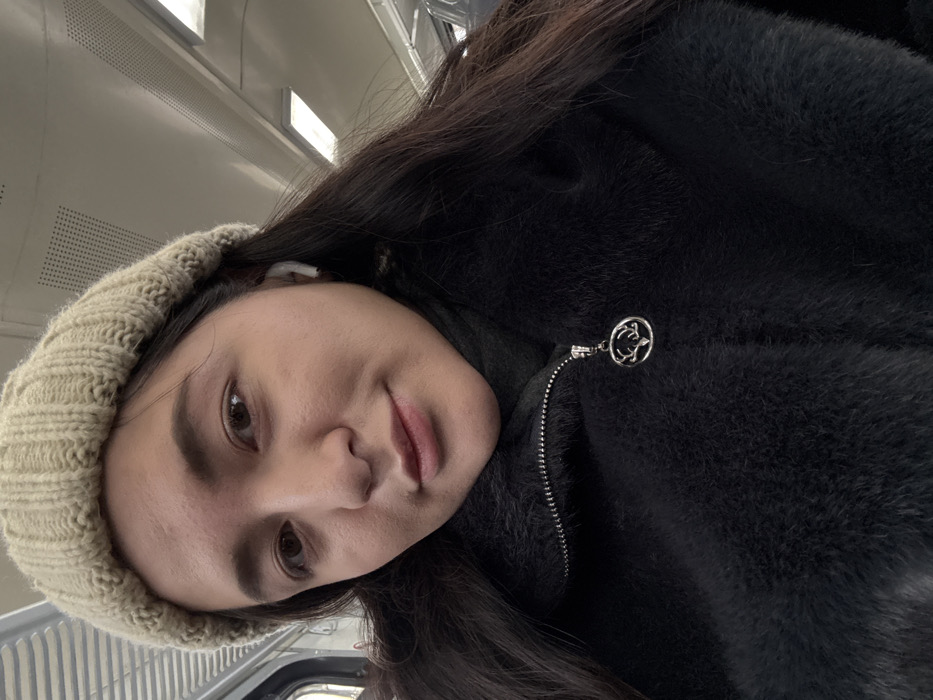 A female individual is depicted in a close-up, self-portrait image. She has long dark hair, a light complexion, and brown eyes, and is looking directly at the camera with a subtle smile. She wears a beige ribbed knit beanie and a black, possibly faux fur, zippered coat over a dark turtleneck. A silver-toned circular pendant featuring a deer's head and antlers hangs from the coat's zipper pull. A white wireless earbud is visible in her right ear. The background shows the interior of a public transport vehicle, characterized by a light-colored ceiling, horizontal fluorescent light fixtures, and a grid-patterned vent. A partial view of a window or door frame with a vertical handle is present on the left. This scene is situated within a public transport vehicle in Il'inskiy Pogost, Russia.