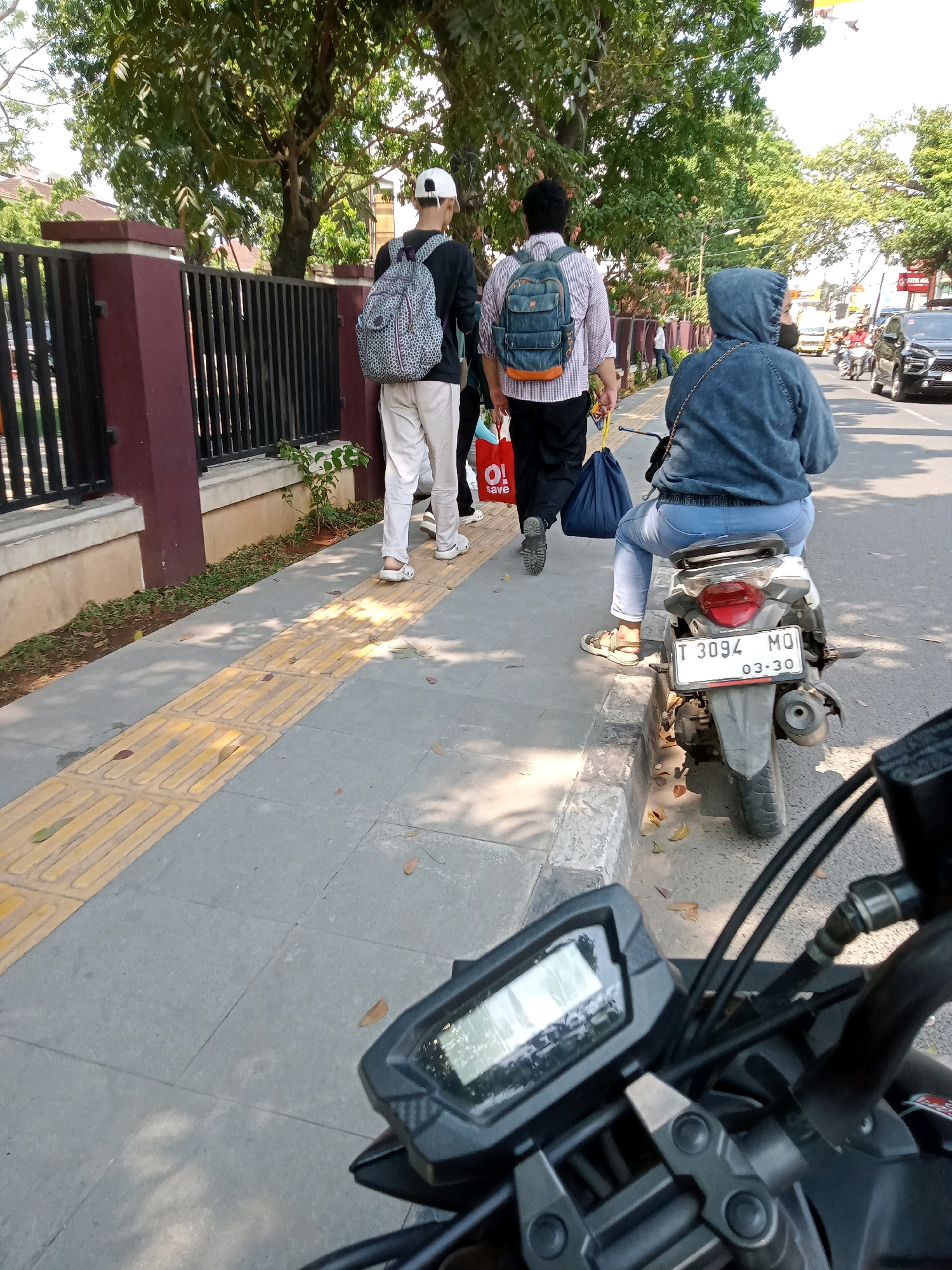 In Karawang, Indonesia, two individuals walk along a sidewalk. The first person, wearing a white cap and a patterned backpack, is dressed in a dark long-sleeved shirt and light-colored pants. Behind them, another person carries a blue backpack and a yellow shopping bag. A woman in a blue hoodie and jeans sits on a motorcycle, facing away from the camera. The motorcycle's license plate reads "T 3094 MQ". The sidewalk is bordered by a dark metal fence and trees. The street is paved, with other vehicles visible in the distance. The time of day appears to be daytime, with sunlight casting shadows.