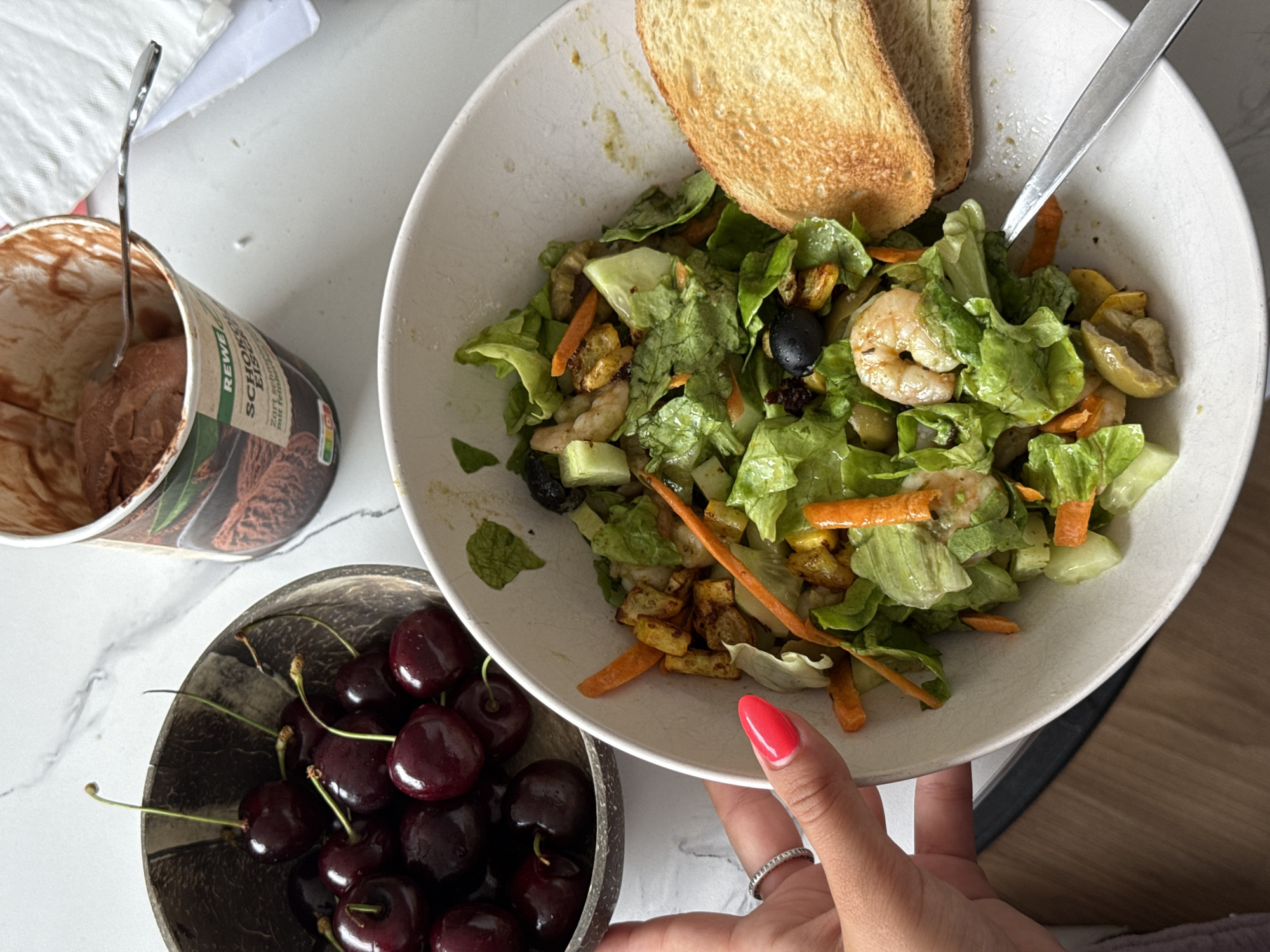 In an indoor setting, possibly a kitchen or dining area, a close-up view shows a collection of food items arranged on a light-colored, speckled countertop. A woman's hand, with bright red nail polish and a ring on her index finger, is visible on the left side of the frame, reaching towards a bowl of dark red cherries. The cherries are served in a textured, dark brown bowl that appears to be made from a coconut shell. To the right of the cherries, a white bowl contains a salad composed of lettuce, sliced carrots, cucumber, olives, and shrimp. Two slices of toasted bread are placed on the edge of the salad bowl. Further to the right, a partially eaten container of chocolate ice cream is visible, with a silver spoon resting inside. The environment suggests a casual meal. This scene is not in Bruchköbel, Germany.