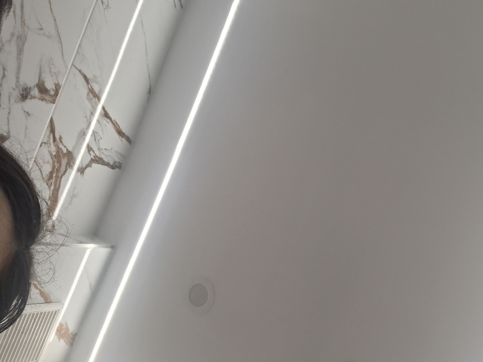 The image depicts an interior scene, likely a bathroom. A person's hair is partially visible on the left side of the frame. To the left, there is a white marble wall with brown veining and white recessed lights. The rest of the scene is dominated by a plain white wall, and there is a circular recessed light fixture near the bottom. The location of the scene in Sofia, Bulgaria, cannot be determined from the image alone.