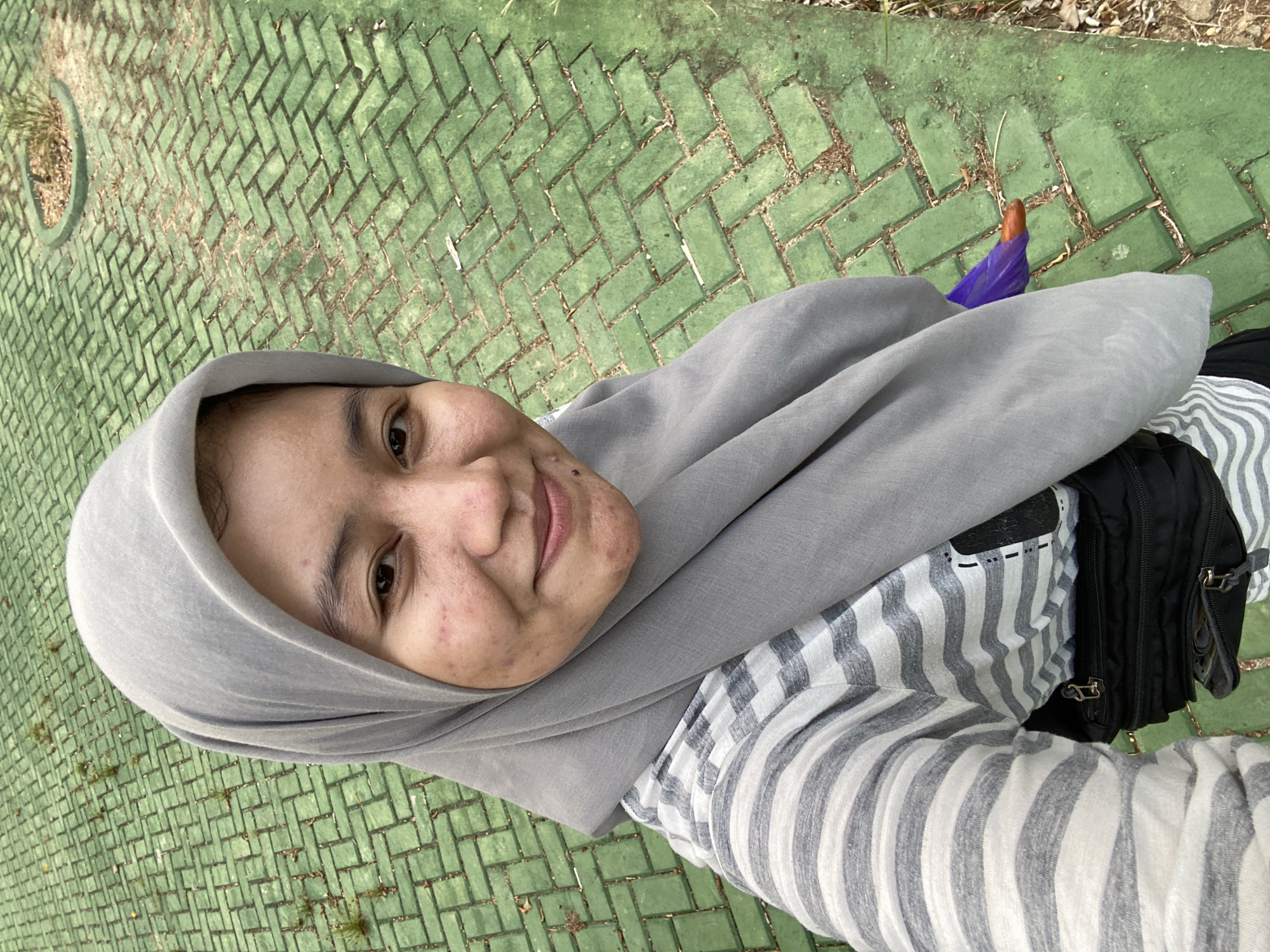 A woman wearing a grey hijab and a striped shirt takes a selfie in front of a green brick pathway. She has a fanny pack around her waist and a purple umbrella peeking out from under her hijab. The pathway is made of rectangular green bricks arranged in a herringbone pattern. There are small plants growing between the bricks. The woman is smiling and looking at the camera. The background is out of focus, but it appears to be an outdoor setting, possibly a park or a public area. The lighting suggests it is daytime.