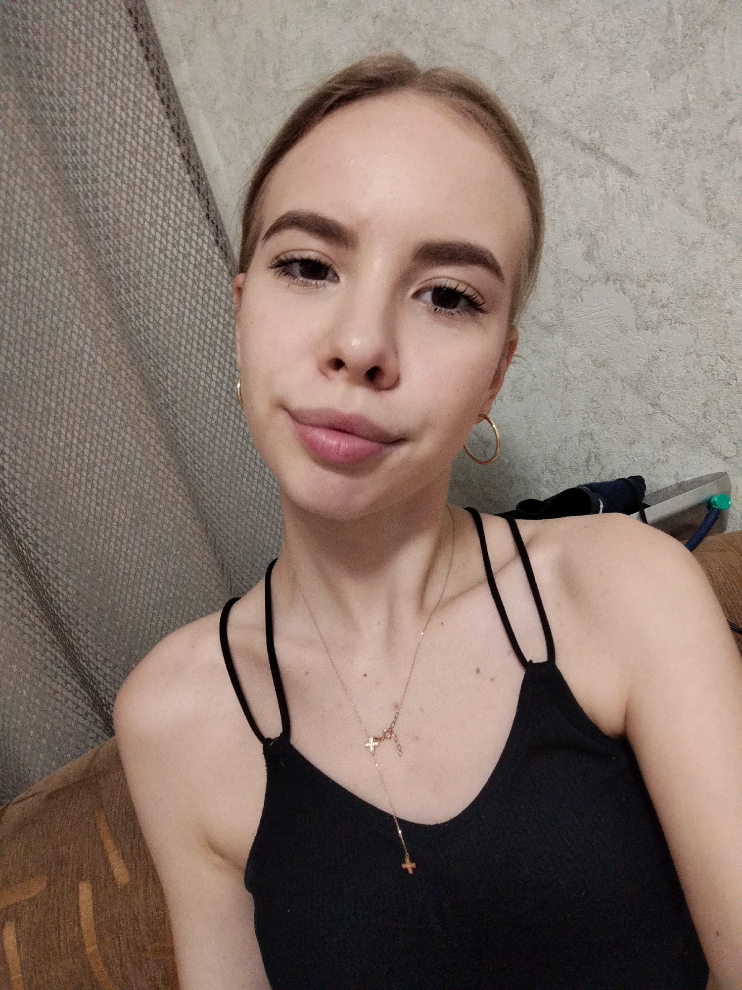 A young woman with blonde hair pulled back, light skin, and dark eyebrows is captured in a close-up, chest-up selfie. She wears a black tank top with thin straps and a delicate necklace featuring two small, gold crosses. Large, gold hoop earrings adorn her ears. Her eyes are dark brown, and her lips are slightly parted. The background is a textured grey wall, with a glimpse of a textured grey curtain on the left and a brown couch and some objects on the right. The lighting appears to be artificial and is casting a soft glow. The scene is indoors, with no specific city or location details discernible.