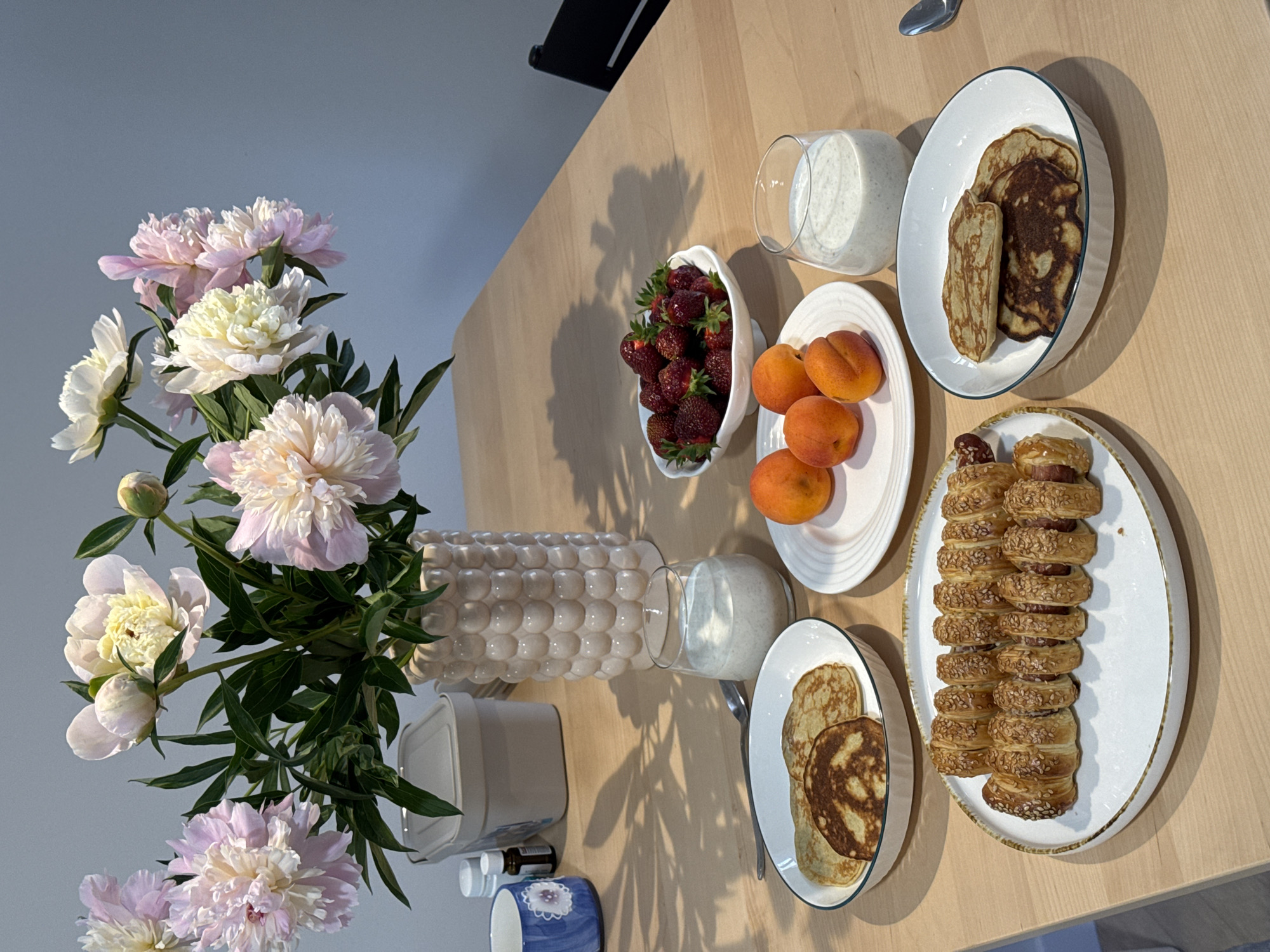 A table is set with a breakfast spread, including pancakes, strawberries, apricots, and two glasses of a creamy beverage. A bouquet of pink and white peonies in a decorative vase sits behind the food. There are also two bottles and a blue mug to the left of the vase. The scene is indoors, likely a home dining area, with a light-colored wooden table and a neutral-toned wall in the background. The lighting suggests it is daytime, with shadows cast by the flowers and vase. There are no people visible in the image. The location of Izhevsk, Russia, is not indicated by any visual cues within the image.