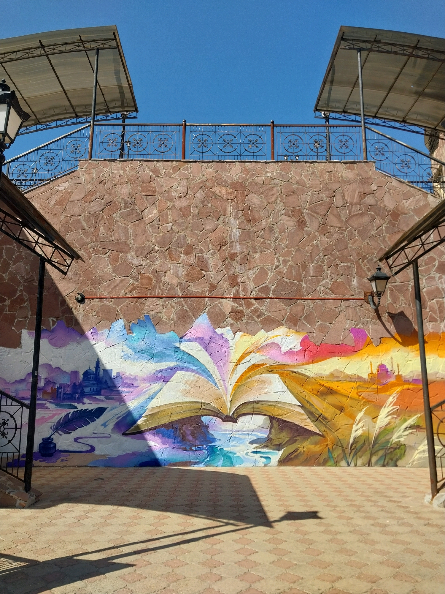 The image shows a vibrant mural painted on a stone-clad wall in Orenburg, Russia. The mural depicts an open book with pages transforming into colorful landscapes, symbolizing knowledge and imagination. On the left side of the mural, a quill pen dipped in an inkwell rests beside a silhouette of a city, possibly representing Orenburg itself. The right side of the mural features a grassy field with tall reeds under a bright, sunlit sky. Above the mural, a stone staircase leads up to a walkway with a wrought-iron railing and two covered sections, suggesting an outdoor seating or viewing area. The ground in front of the mural is paved with interlocking bricks, and shadows cast by overhead structures create a geometric pattern. The overall scene is brightly lit by the sun, indicating a clear day.