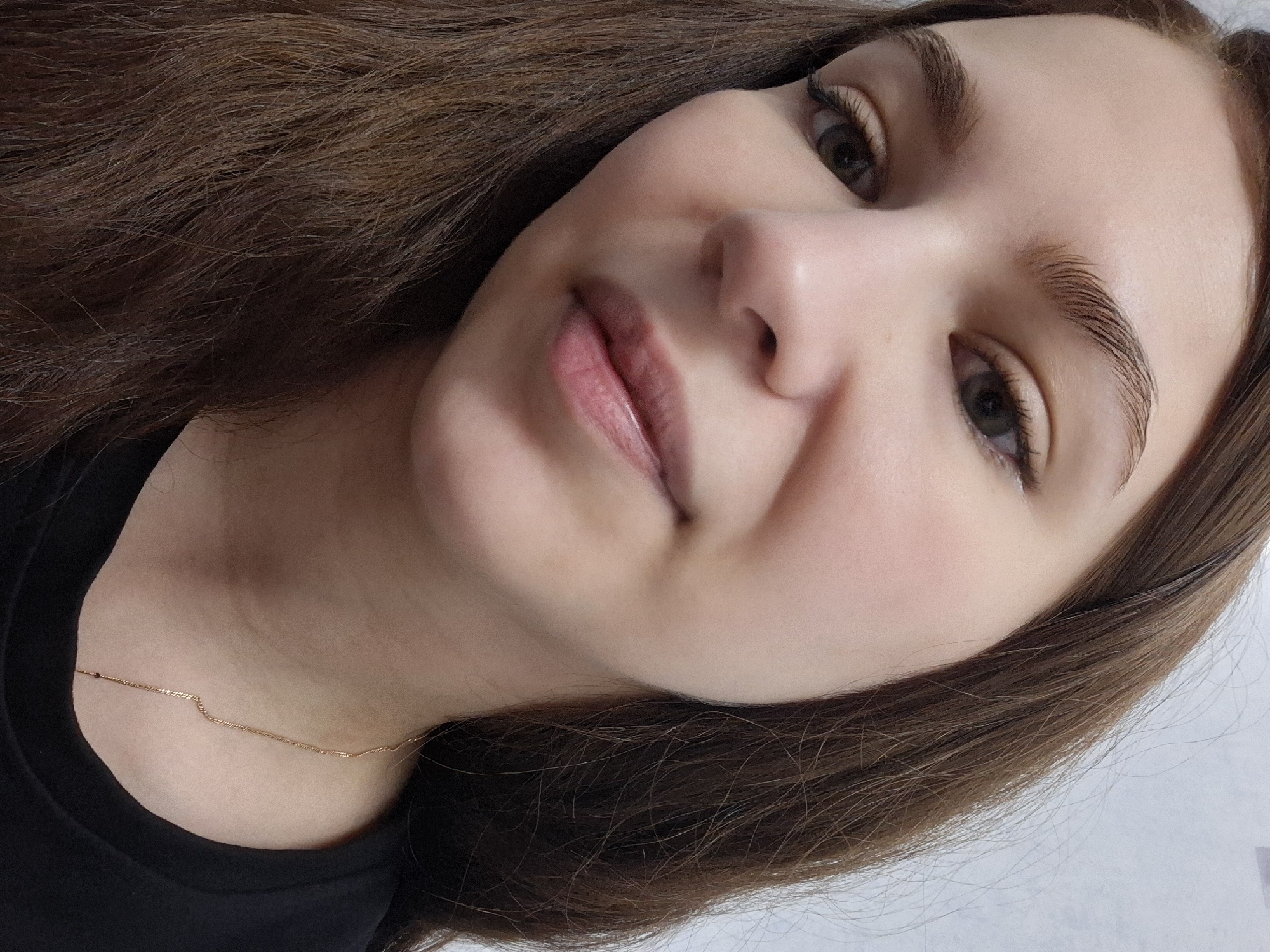 A close-up portrait shows a young woman with long, wavy brown hair, cascading down her left side and partially visible on her right. Her face is angled slightly upwards and to the right, with her gaze directed towards the camera. She has fair skin with a soft blush on her cheeks, dark eyes with visible lashes enhanced by mascara, and meticulously groomed eyebrows, styled upwards. Her lips are a natural, glossy pink, slightly parted. She appears to be wearing minimal makeup, highlighting her natural features. A thin, golden chain necklace rests on her collarbone, partially visible above a simple black top or t-shirt. The background is a plain, light color, suggesting an indoor setting. The lighting is even and soft. The woman's expression is calm and neutral, with a hint of a gentle smile. There is no visible text in the image. The image is taken in Chaikovsky, Russia.