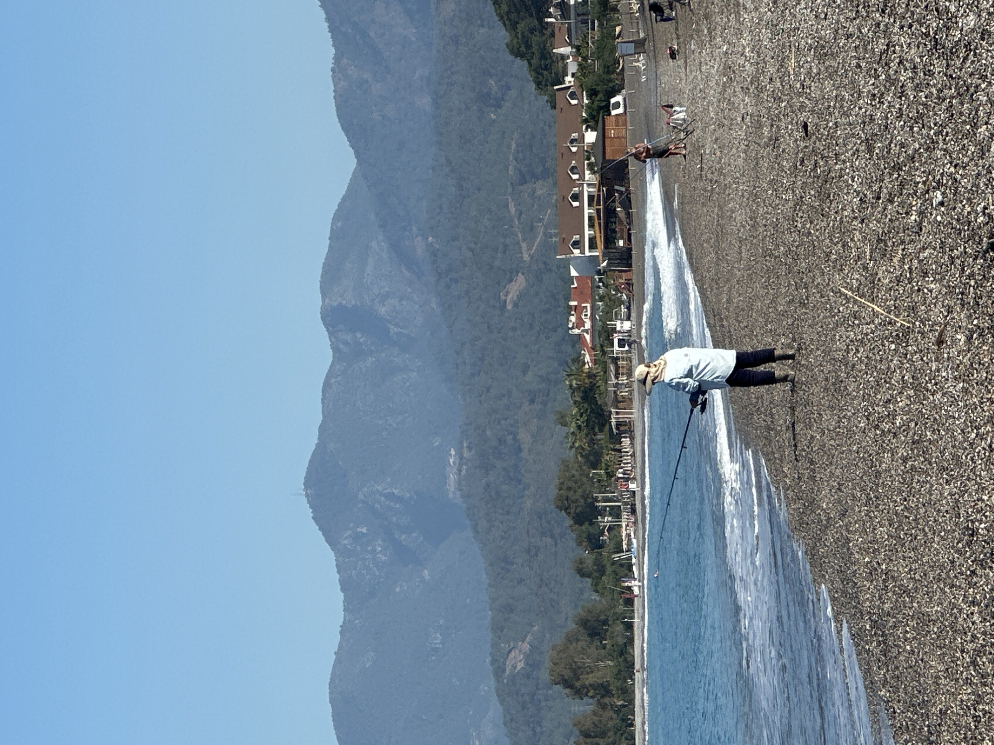 The image depicts a beach scene in Fethiye, Turkey. A person wearing dark pants, a light blue shirt, and a hat stands on the pebble beach near the water's edge, holding a fishing rod. Buildings and trees line the coast behind them, and mountains are visible in the background under a clear blue sky. Other people can be seen further down the beach.