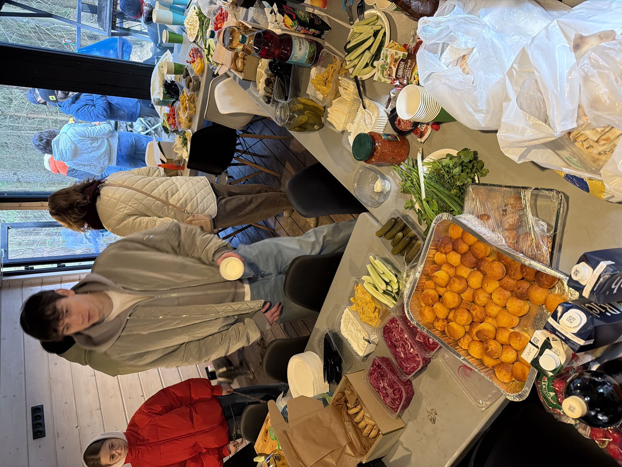 This image depicts a casual indoor gathering around a long table laden with a buffet-style spread of food and drinks. The setting appears to be a modern cabin or building with large windows overlooking a wooded area, suggesting a natural or rustic environment like Vsevolozhsk, Russia.
Several people are present, dressed in warm clothing, indicating cool weather. One person in a white quilted jacket stands by the table, while another in a light grey jacket and light blue jeans holds a white cup. In the foreground, a person wears a bright red puffer jacket and a white hoodie. More individuals in winter jackets and hats can be seen in the background, some reflected in the window. The bare trees outside reinforce the impression of autumn or winter.
The table is filled with various items: a large aluminum tray holds numerous small, golden-brown baked spheres (possibly potato balls). Other dishes include containers of sliced fresh cucumbers, pickled cucumbers, and red pickled cabbage. There are also bowls of chips or nachos, a creamy dip, fresh herbs (dill, green onions, cilantro), and a jar of red sauce. Drinks include bottles of juice (one visibly labeled "Черноголовка" - Chernogolovka), "Rich" brand juice cartons, and a wine bottle. Disposable paper cups, plates, and black plastic cutlery are set out for use. Several white plastic bags, likely containing bread or flatbreads, are piled at one end, and a cardboard box holds cookies or biscuits.
The activity is a social event, likely a meal or party, where guests are serving themselves from the communal food setup. The scene suggests a relaxed and informal atmosphere. Visible text includes "ЧЕРНОГОВКА" (Chernogolovka) on a bottle, "СЛАБОДА" (Sloboda) on a snack package, and "RICH" on juice cartons, all in Cyrillic.