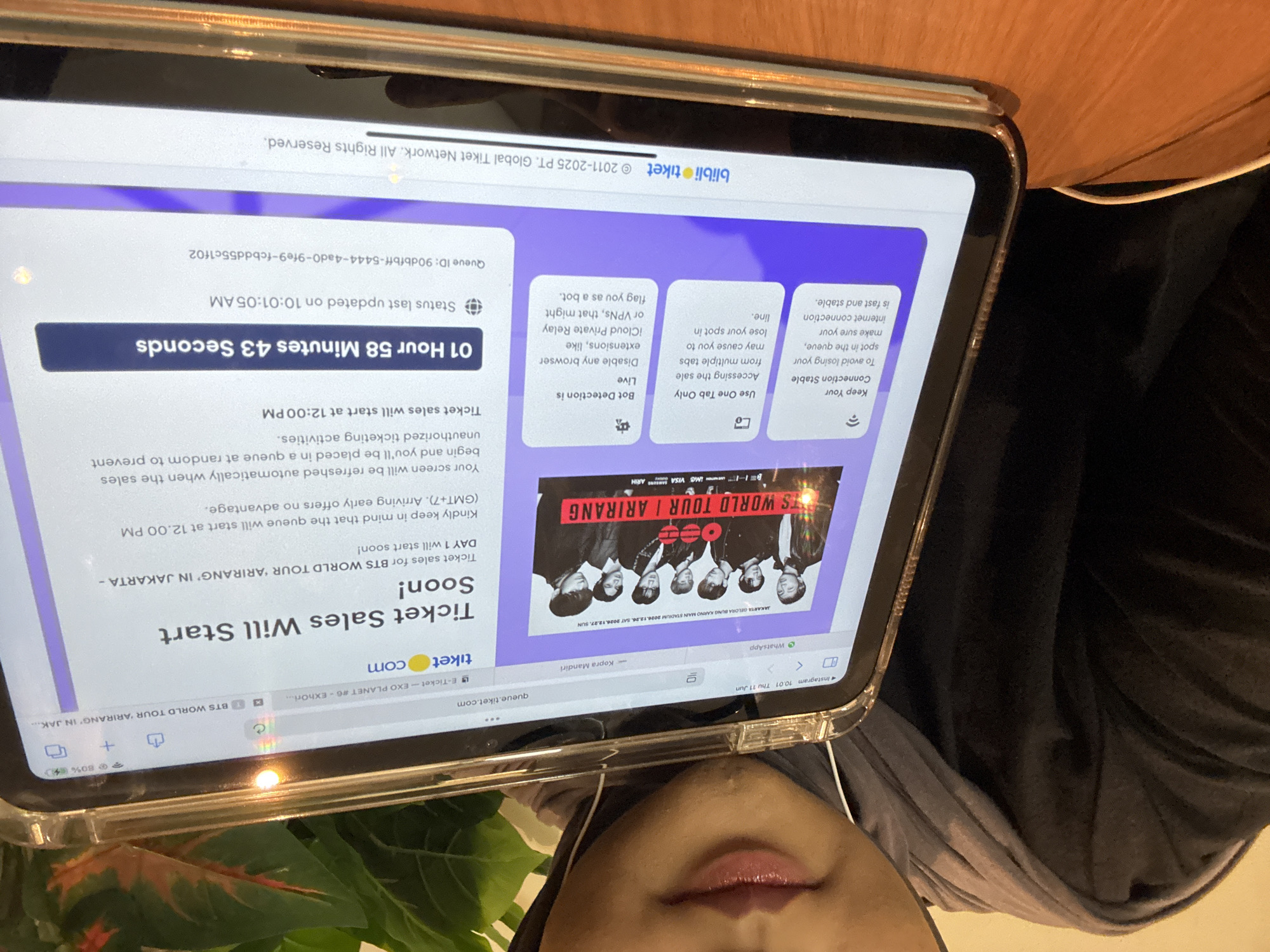An overhead shot shows a person's face and shoulders, wearing a grey hijab and hoodie, as they hold a tablet computer in landscape orientation. The tablet displays a webpage with information about BTS ticket sales for the "WORLD TOUR | ARIRANG" in Jakarta. The webpage indicates that ticket sales will start soon, with the queue opening at 12:00 PM GMT+7. A countdown timer shows "01 Hour 58 Minutes 43 Seconds" remaining. The website footer shows the blibli tiket logo and copyright information. The tablet is placed on a wooden surface, possibly a table. In the background, a green plant with reddish-orange leaves is visible, along with parts of a dark clothing item. The lighting appears to be artificial, with a noticeable glare on the tablet screen. The location is identified as West Jakarta, Indonesia, based on the text on the webpage which specifies "JAKARTA GELORA BUNGO KARNO MAIN STADIUM 2026.12.24, SAT 2026.12.27, SUN".