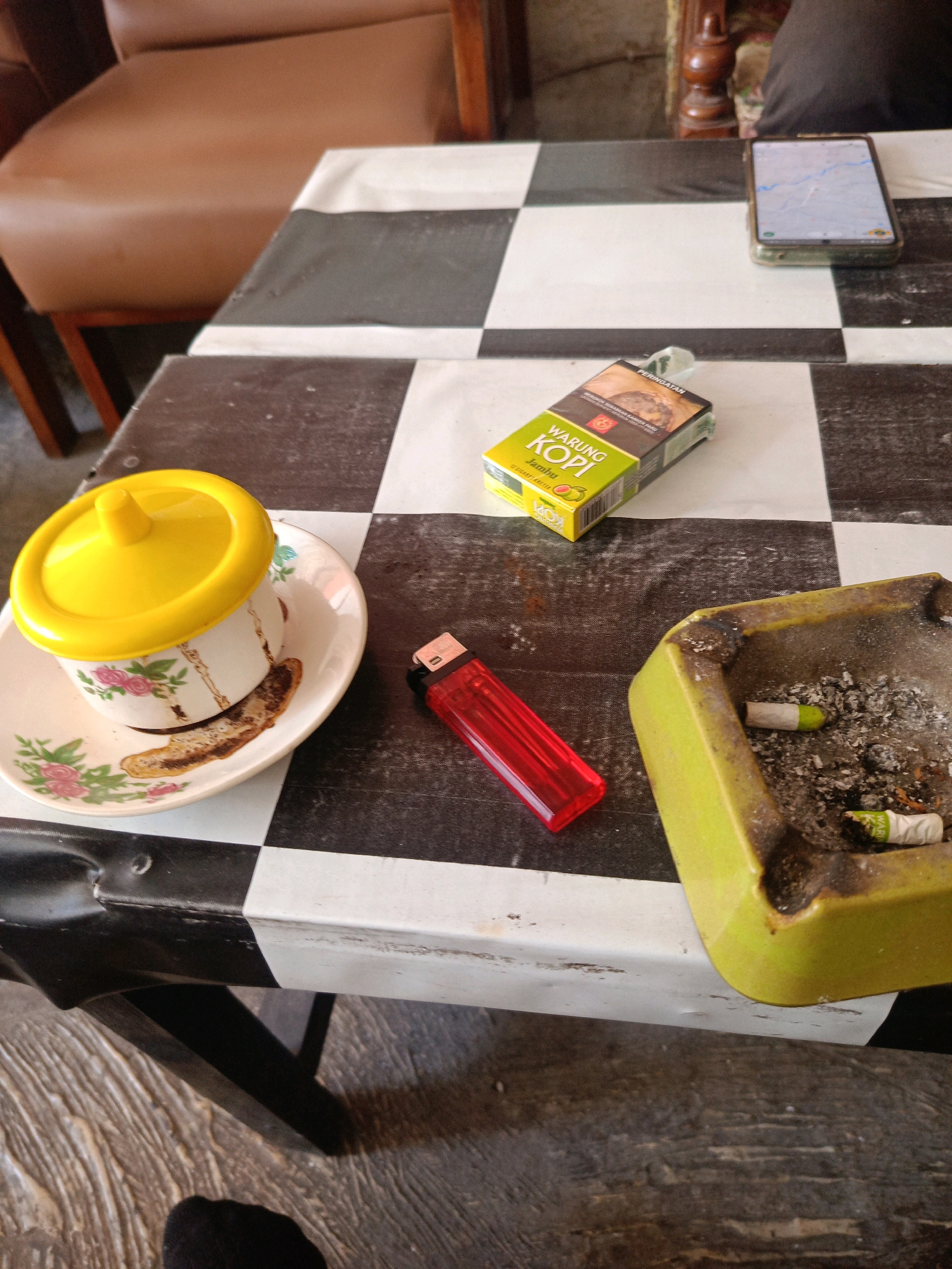 A close-up, high-angle shot captures a table with a black and white checkered cloth, likely in Candirejo, Indonesia. On the table rests a light brown armchair and a portion of a wooden chair. The foreground features a white ceramic cup with a floral pattern, a yellow lid, and a saucer stained with coffee residue. Adjacent to the cup is a red lighter. Further to the right, a bright yellow ashtray holds several cigarette butts and ashes. A pack of "WARUNG KOPI Jambu" cigarettes is placed near the center of the frame, and a smartphone displaying a map application is visible in the background. The scene suggests a moment of pause or relaxation, possibly at a cafe or home.