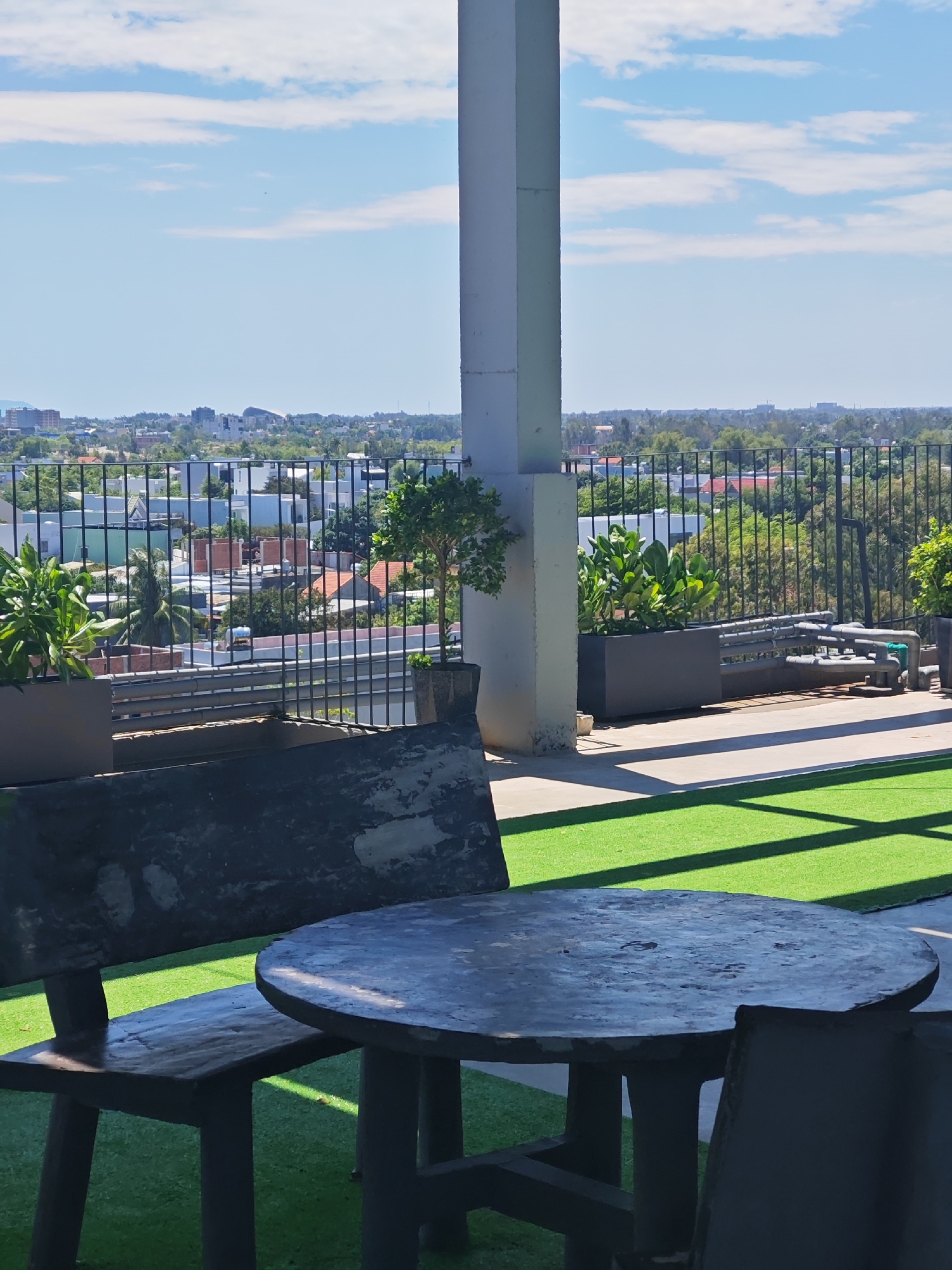 The image depicts a rooftop seating area with a panoramic view of Da Nang, Vietnam. In the foreground, a dark-colored, weathered bench and a round table are set on bright green artificial turf. Behind them, a tall, light-colored concrete pillar supports an overhead structure, casting shadows on the turf. A black metal fence runs across the middle ground, separating the seating area from the cityscape. Beyond the fence, a dense residential area with a mix of white and orange-roofed houses and lush green trees stretches towards the horizon. In the distance, taller buildings and a distinctive curved structure are visible against a pale blue sky with scattered white clouds. To the left of the pillar, a small, potted tree with dark green foliage stands in a dark planter box. To the right of the pillar, another dark planter box holds a cluster of large, leafy green plants. A section of pipes and valves is visible behind the planter box on the right. The scene suggests a relaxed outdoor space offering a commanding view of the city.