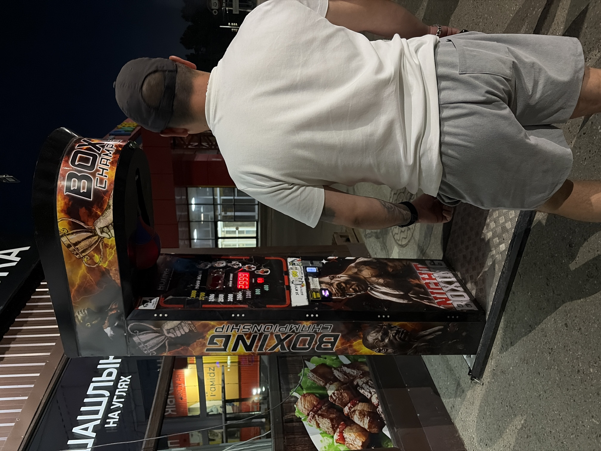 The image shows a man standing in front of a boxing arcade game. The game is decorated with images of boxers and a trophy. The man is wearing a white t-shirt and gray shorts. He has a baseball cap on his head and tattoos on his arm. The boxing game has a digital display showing a high score of 2999. In the background, there is a sign with Russian text that translates to "Shashlik on the corners". The scene appears to be outdoors at night, possibly in a city park or a street market. The man is looking at the boxing game, and it is unclear if he is playing it or about to play it. The overall impression is that of a casual evening entertainment setting.