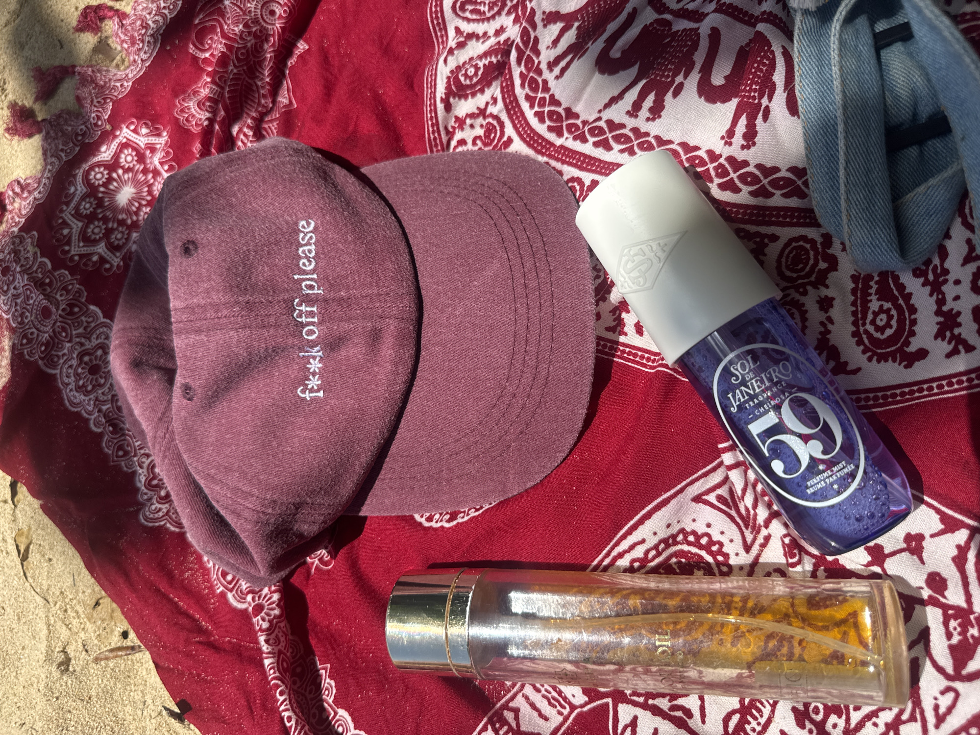 The image shows a flat lay of personal items on a red bandana with a paisley pattern, placed on sand. In the center is a maroon baseball cap with the embroidered text "f**k off please." To the left of the cap is a clear glass bottle containing a yellow liquid, likely perfume or oil, with a gold cap. To the right of the cap is a purple bottle of "SOL DE JANEIRO" mist. A light blue denim fabric, possibly from jeans, is visible in the bottom right corner. The scene is illuminated by natural sunlight, casting shadows on the objects and creating a warm, summery atmosphere. The location is identified as Choeng Thale, Thailand.