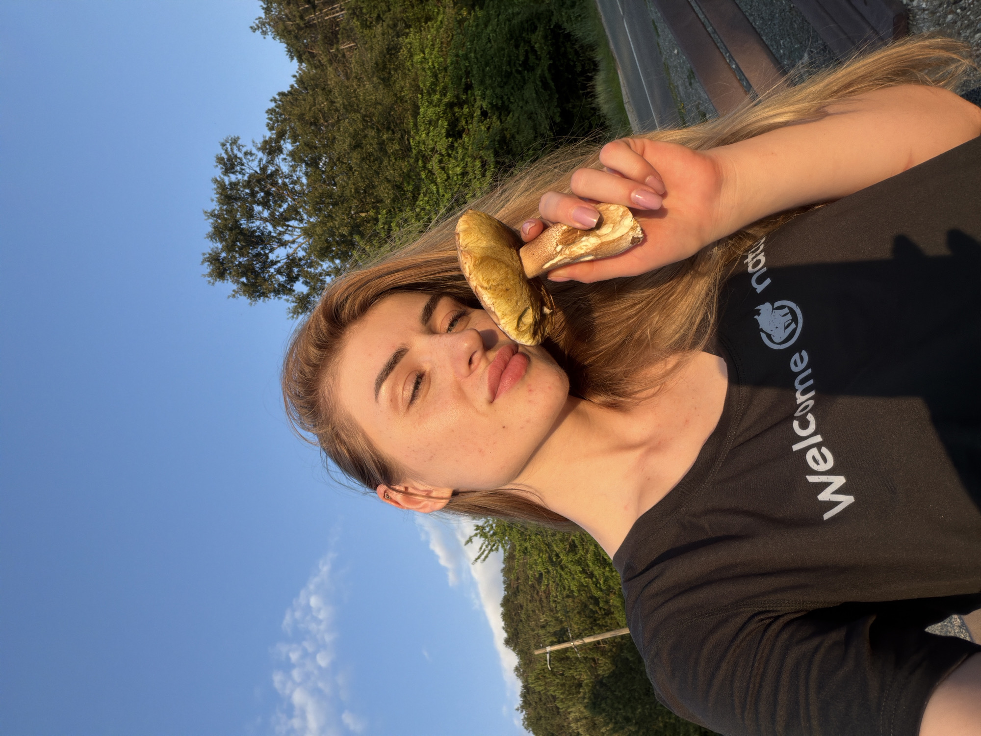 A young woman is shown in a selfie holding a large mushroom to her face. She has long blonde hair and is wearing a black t-shirt with white text and a logo. Her eyes are closed, and she is smiling. She is outdoors, with a blue sky and trees in the background. The sun is setting, casting a warm glow on her face. The scene appears to be in a rural or wooded area, possibly a park or nature reserve. The woman is looking at the camera, and the mushroom is held close to her lips. The overall impression is one of happiness and connection with nature.