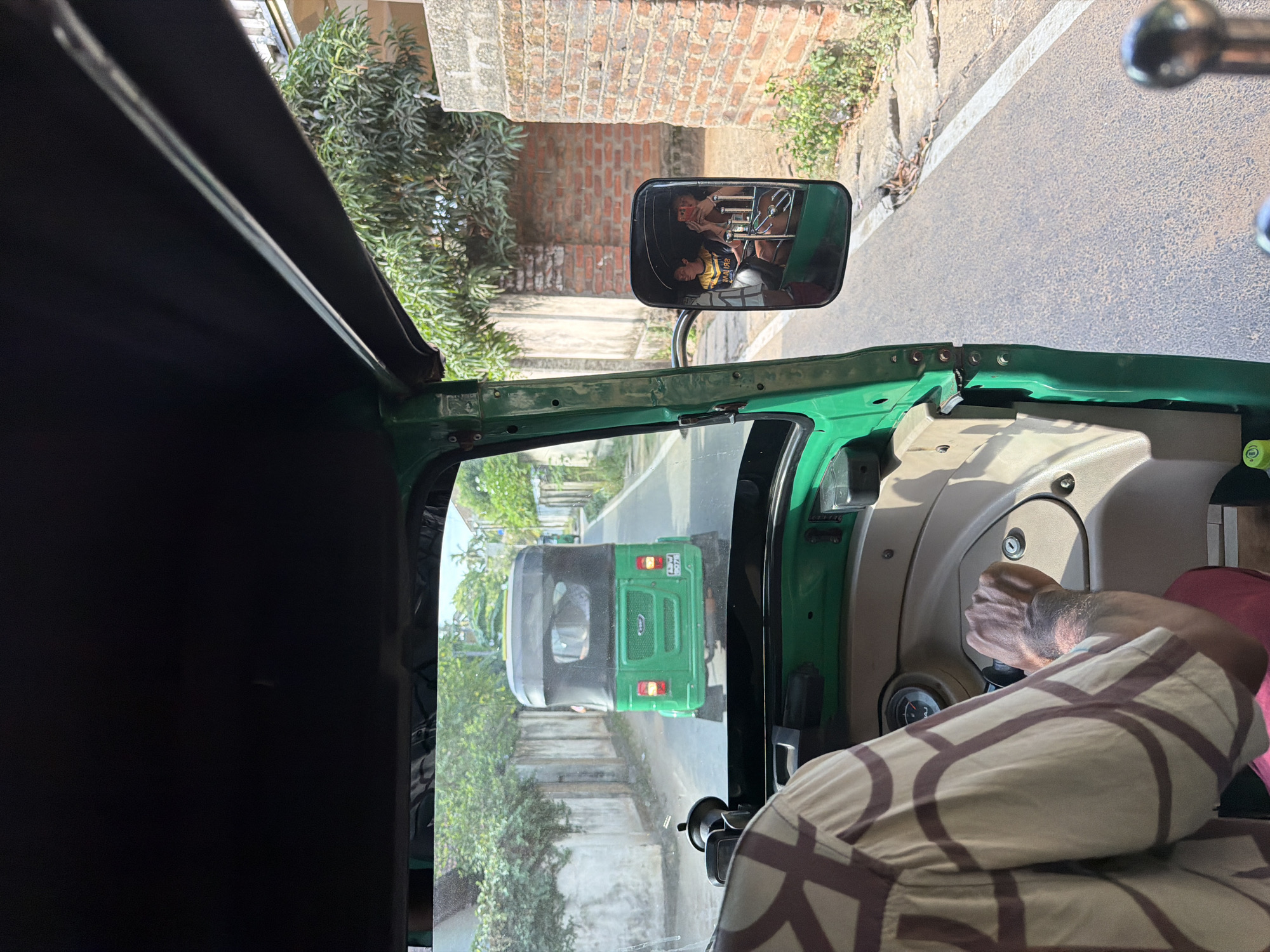 The image captures a view from inside a green auto-rickshaw, looking forward and to the right. The driver's left arm, wearing a patterned shirt, is visible gripping the steering wheel. The dashboard of the auto-rickshaw is visible, including a gauge and part of the steering column. In the foreground, a side mirror reflects two individuals, one holding a smartphone, presumably passengers within the auto-rickshaw.
The auto-rickshaw is traveling on a paved road, indicated by the white line on the right side. On the left side of the road, there is a low, weathered stone wall, with trees and foliage growing beyond it. On the right side of the road, there is a brick wall and more dense foliage. Ahead, another green auto-rickshaw is visible on the road, also traveling in the same direction. The environment appears to be a semi-urban or rural setting with evident signs of vegetation and simple structures. The lighting suggests it is daytime with bright sunlight casting shadows. This scene is consistent with the roads and typical modes of transport found in Pottuvil, Sri Lanka.