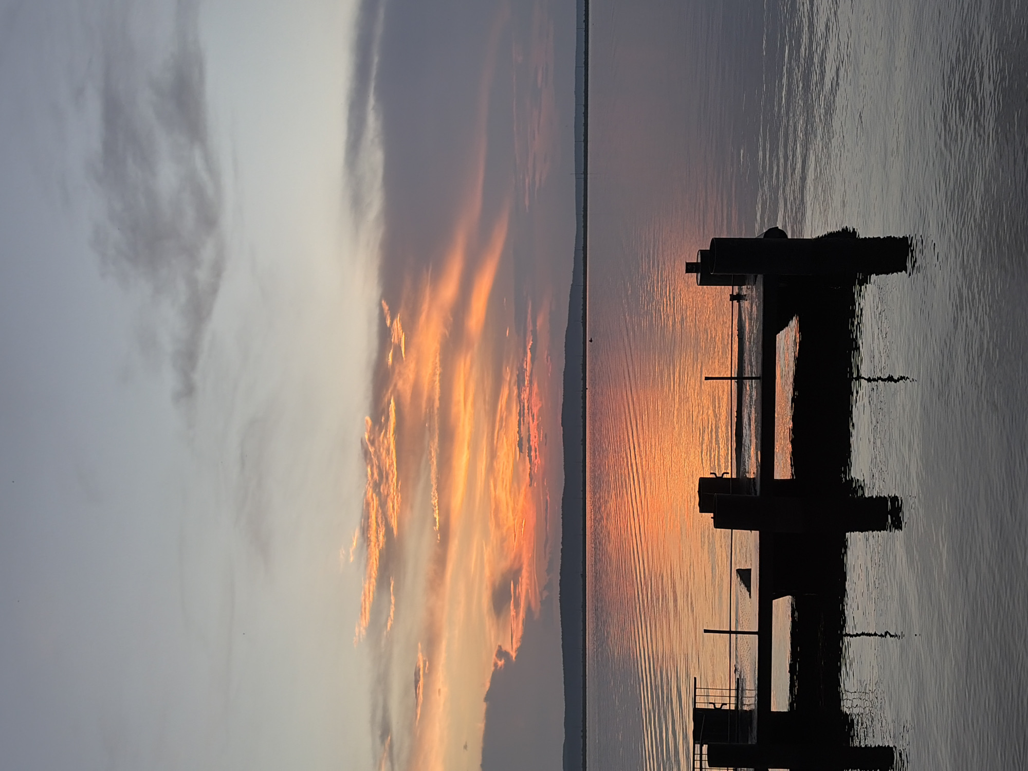 A sunset over the Kama River in Chaikovsky, Russia, is captured in this image. The sky is painted with vibrant hues of orange and pink, with streaks of darker clouds adding depth and contrast. The sun, low on the horizon, casts a warm, golden reflection onto the rippling water. In the foreground, a silhouetted dock with thick pilings juts out into the river. The water's surface is calm, reflecting the colors of the sky and the dark structure of the dock. The far bank of the river is a dark, mountainous silhouette, adding a sense of vastness to the scene. The overall atmosphere is serene and tranquil, evoking the beauty of nature at dusk.