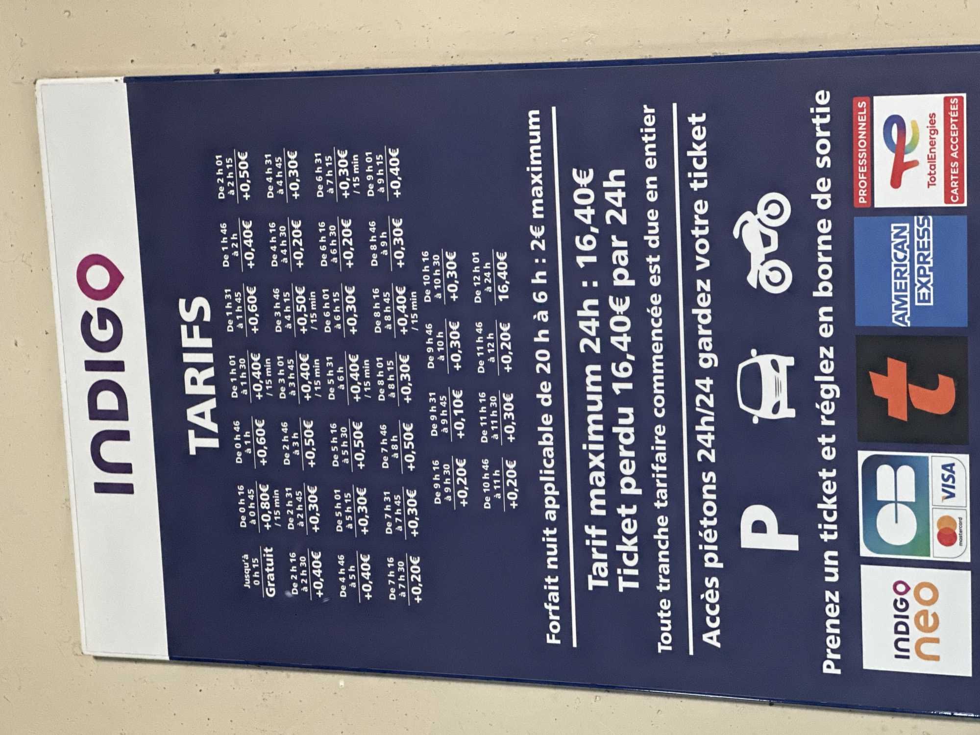 This is a photograph of a parking rate sign from Indigo Neo in Dieppe, France. The sign displays various hourly parking rates, a night tariff, a maximum 24-hour charge, and instructions for pedestrian access and payment. The Indigo Neo logo is prominently displayed. Below the main pricing information, there are logos for various payment methods including CB, Visa, Mastercard, American Express, and TotalEnergies cards. The sign appears to be mounted on a wall or a similar vertical surface in a public parking area. There are no people or actions visible in the image, only the static display of the pricing information.