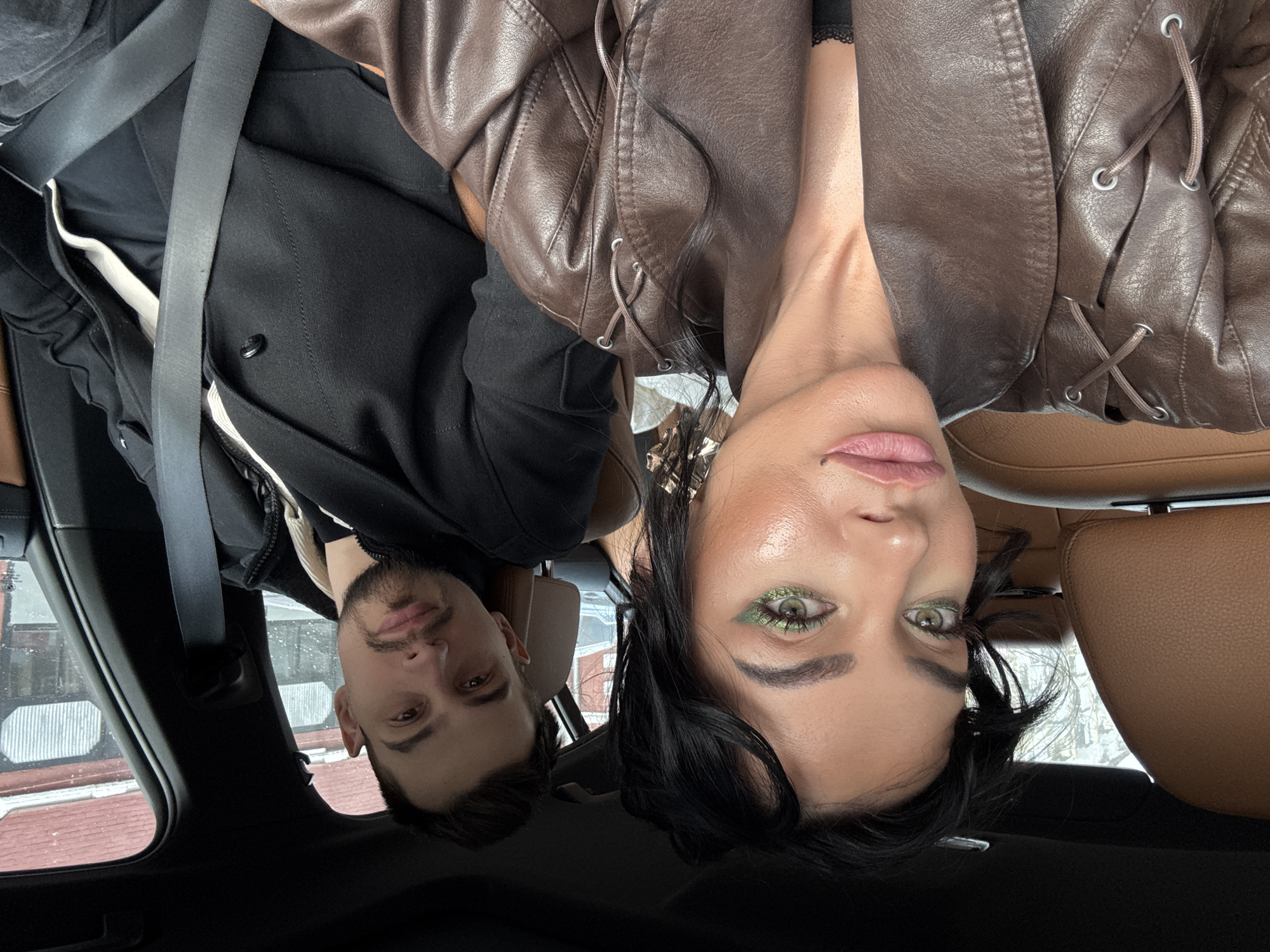 An image captures two individuals, a man and a woman, looking towards the camera from inside a car. The woman is in the foreground, wearing a brown leather-like jacket with decorative lacing details on the shoulders. She has dark hair, green shimmering eyeshadow, and light pink lipstick, with a small mole on her upper lip. The man is positioned slightly behind her, with dark hair and a beard, wearing a dark jacket or hoodie. Both have neutral expressions.
The car's interior is visible, featuring brown leather seats and dark trim. A black seatbelt crosses the man's chest. Through the car windows, a wintery outdoor scene is apparent, with snow covering structures and the ground, and bare trees. Buildings with red and corrugated metal roofs are visible in the background, suggesting an urban or suburban setting. The scene takes place during the daytime. The location context provided is Krasnoyarsk, Russia. No visible text appears in the image.