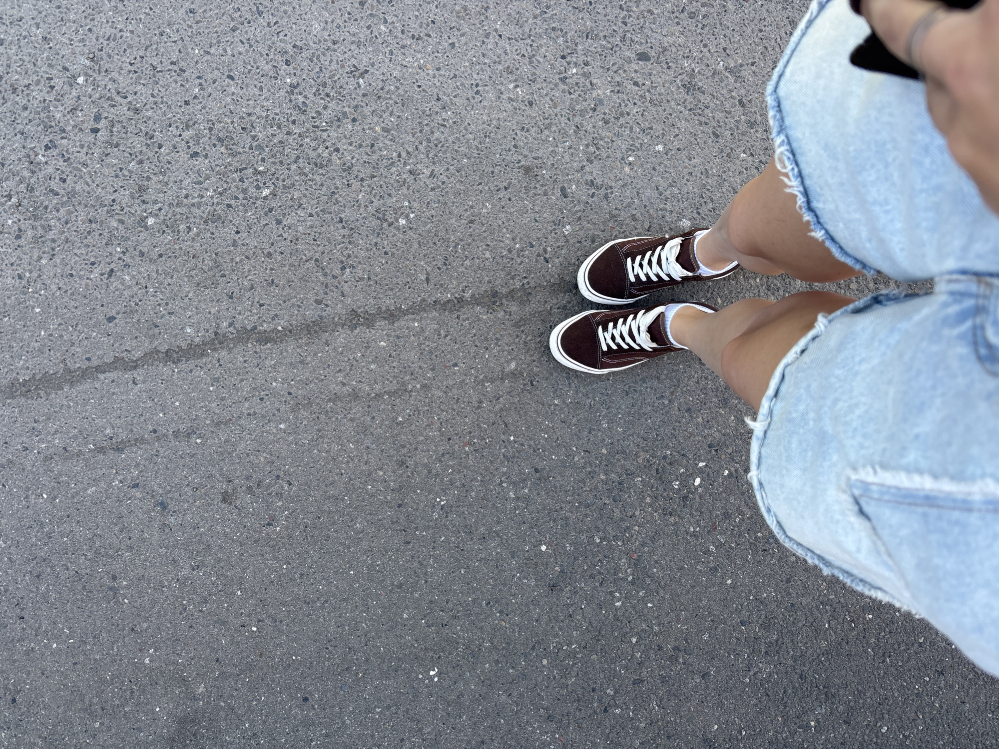 This is an overhead, eye-level shot of a person's legs and feet standing on a textured asphalt surface. The person is wearing dark brown low-top sneakers with white laces and white socks, and a light blue denim skirt with a frayed hem. The skirt occupies the right side of the frame, and the sneakers are positioned on the lower right. The asphalt is dark gray and granular, with a faint line running diagonally across the left side of the image. The lighting suggests it is daytime, and the environment appears to be outdoors on a paved surface, possibly a sidewalk or road. No specific time of day, weather conditions, or strong emotional cues are discernible. There is no visible text in the image.