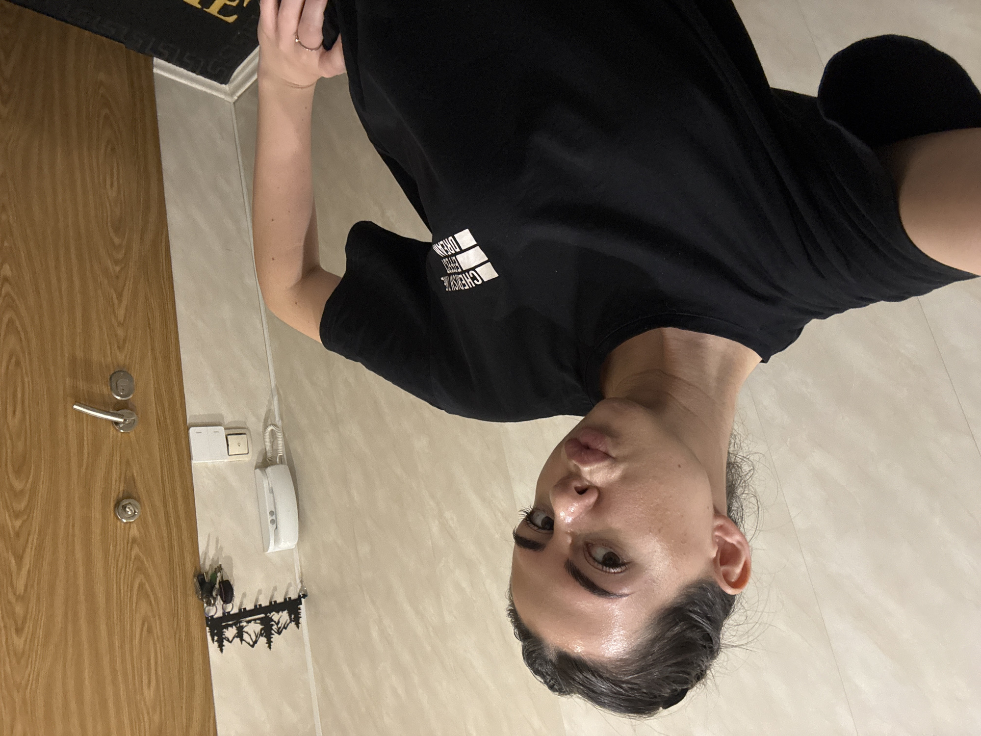 The image shows an individual, appearing to be a young adult female, posing indoors. She has dark hair, pulled back, and is wearing a plain black short-sleeved t-shirt with a white graphic design on the chest. A ring is visible on one of her fingers. She looks directly at the camera with a neutral to slightly serious expression.
The background suggests an entryway or hallway. To her left is a light brown wooden door equipped with a silver handle and lock. On the adjacent wall, a white wall-mounted intercom device and two white light switches are visible. Below them, a black metal key holder, designed with a forest silhouette, hangs with several keys. The walls and floor are covered in light-colored tiles, or similar material, providing a clean, bright backdrop. A dark mat is partially visible on the floor near the door.
The main activity is the person posing for a photograph. The lighting indicates an indoor setting, likely under artificial illumination. On the t-shirt, white text can be seen, partially spelling "CHERYSHE" (or "CHERISH THE") followed by "DREAM", alongside three distinctive white rectangular shapes.