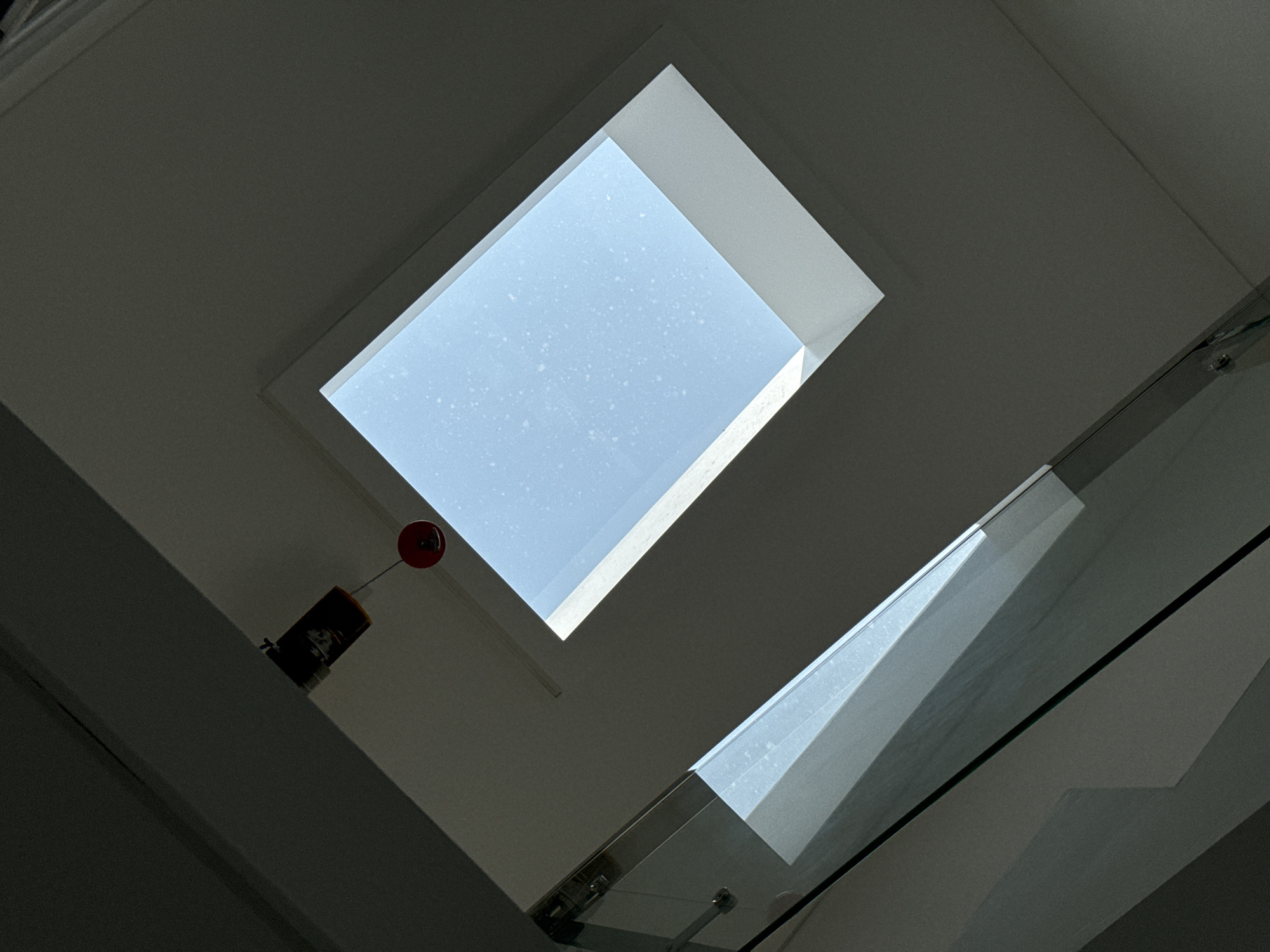 The image shows an interior view looking upwards towards a skylight. The skylight is rectangular and angled, revealing a light blue sky with small white speckles, possibly dust or condensation on the glass. Below the skylight, a dark ceiling angles downwards, creating a stark contrast with the illuminated sky.
In the lower left of the frame, a glass railing of a staircase is visible, reflecting the light from the skylight and the surrounding interior. The glass has a metallic frame. A dark, possibly wooden, beam runs diagonally across the upper left portion of the image, obscuring part of the ceiling. Attached to this beam, and hanging down, is a small, dark rectangular object with some orange and white markings, and below it, a small red round object attached by a thin wire.
The image does not contain any discernible elements that would specifically place it in Tanjungmeru, Indonesia, or any other city. The scene appears to be a modern interior space, possibly a home or a commercial building. There are no people, actions, or interactions depicted.