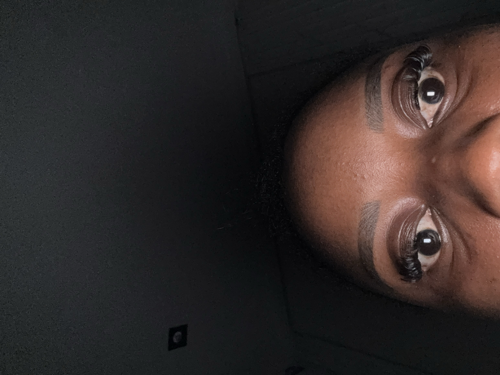 This is a close-up, low-angle, vertical shot of a person's eyes and forehead, against a dark, textured background. The subject's skin is dark brown, and their eyebrows are neatly shaped and filled in. They are wearing false eyelashes that are long and voluminous. The eyes are open, with dark irises reflecting a bright light source, creating small circular highlights. The forehead shows some texture and is lightly creased. A small, square, recessed light fixture is visible in the upper left quadrant of the image, embedded in the dark background. The lighting is dim, with the primary light source illuminating the subject's face from above and slightly in front. The overall impression is of a selfie taken in a dimly lit interior space, possibly a room in Abuja, Nigeria.