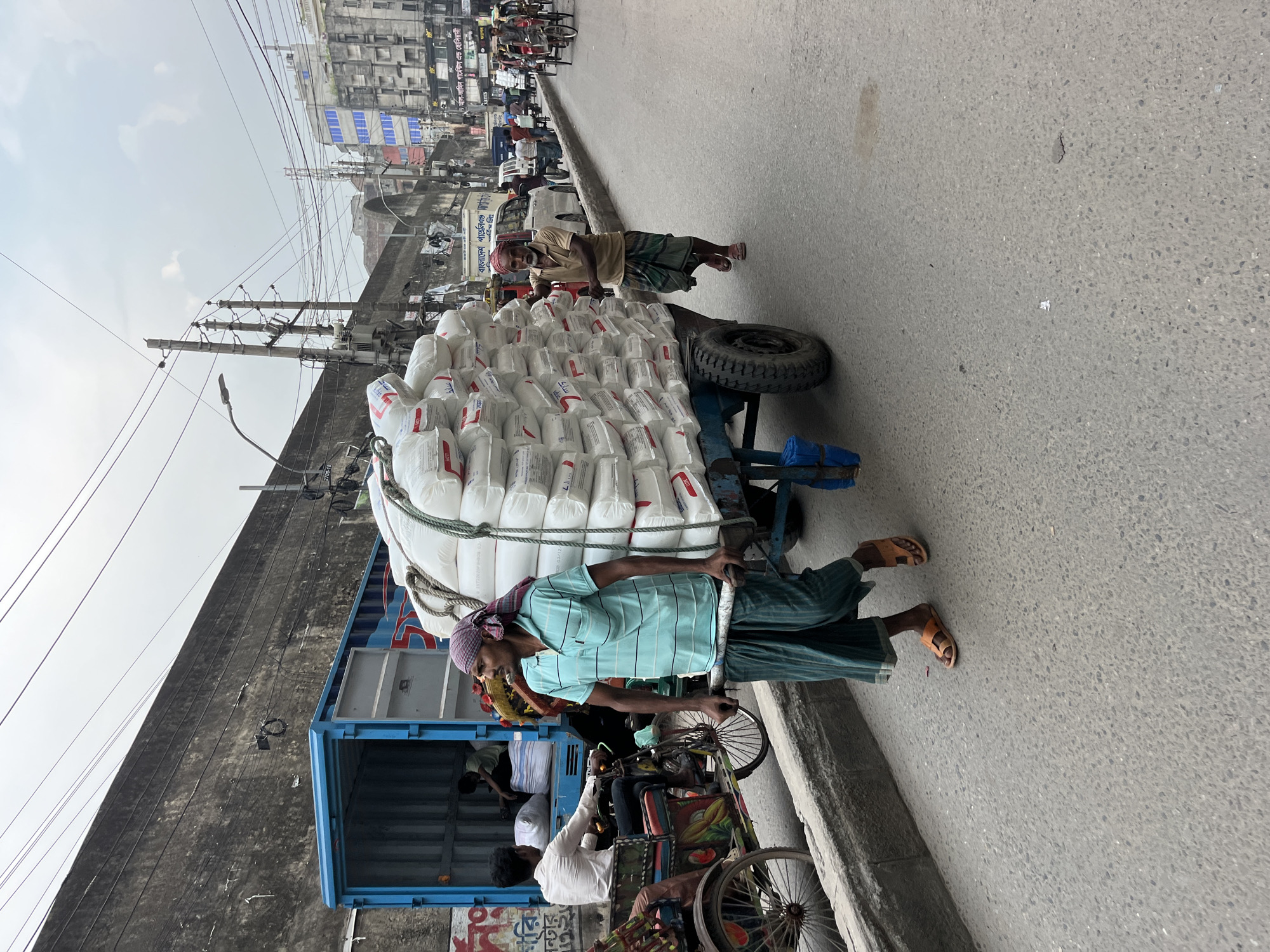 On a street in Dhaka, Bangladesh, a man pulls a two-wheeled cart piled high with numerous white sacks. The cart is overloaded, with the sacks tied together with rope. The man wears a light blue striped shirt, green and white striped pants, a red headscarf, and sandals. Another man is walking beside the cart. Behind the cart, a blue vehicle with an open back is parked next to a three-wheeled bicycle rickshaw. Several people are in or around the rickshaw, while more people can be seen in the background, along with buildings, overhead wires, and other vehicles. The environment indicates a busy street scene.