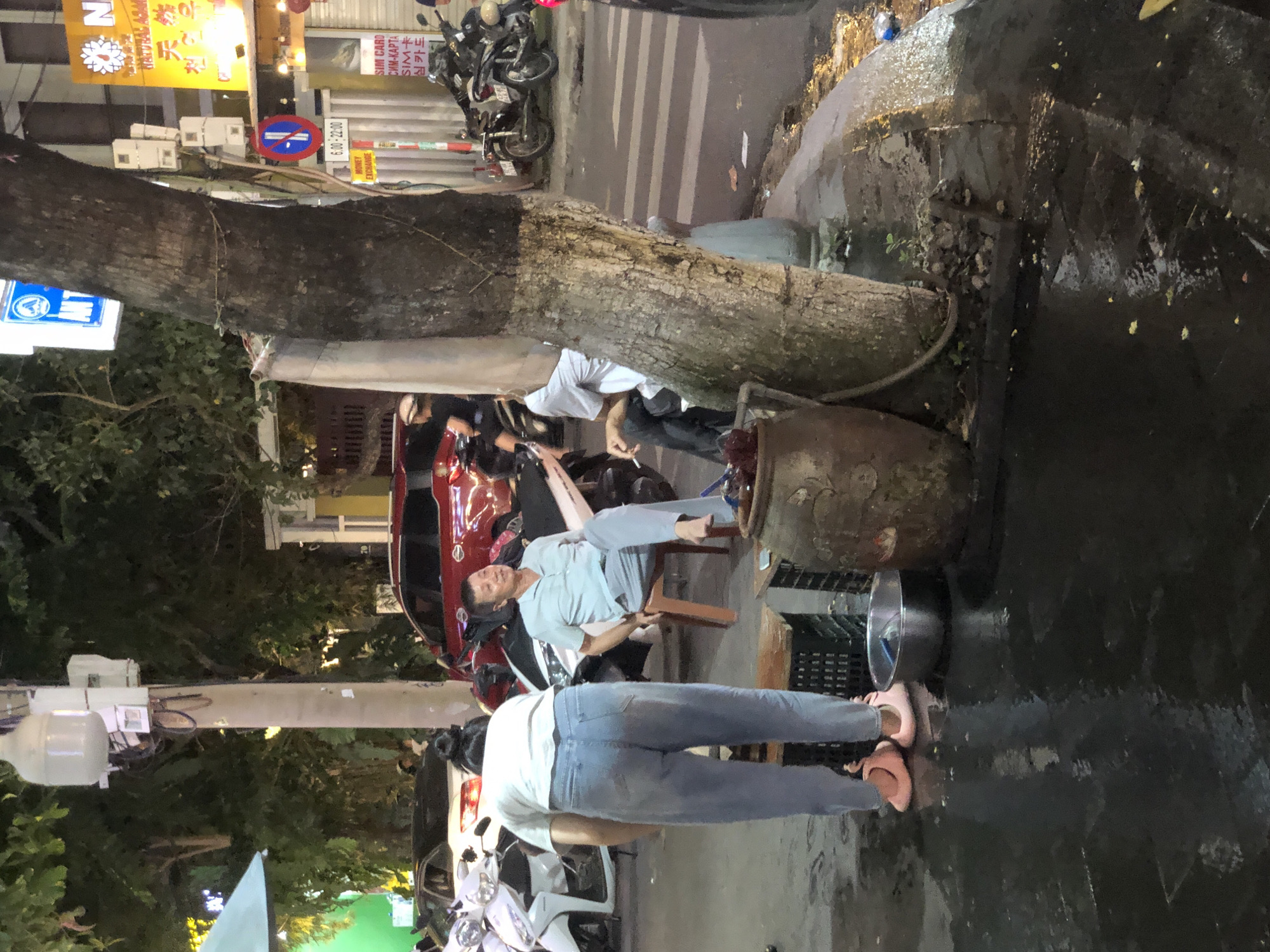 The image depicts a street scene at night in Da Nang, Vietnam. In the foreground, a person wearing light blue jeans and pink flat shoes is bent over, facing away from the camera. To their right, a metal bowl sits on the wet pavement, next to a large, brown ceramic pot. Further back, a man in a light blue short-sleeved shirt and grey pants sits on a wooden stool, looking to his left and smiling. He is seated in front of a motorcycle. Behind him, a red SUV is parked. In the background, trees and streetlights are visible, along with several other motorcycles and the facades of buildings. A no-parking sign and a money exchange sign are posted on a pole near a large tree on the right side of the image. The ground is wet, indicating recent rain or street cleaning.