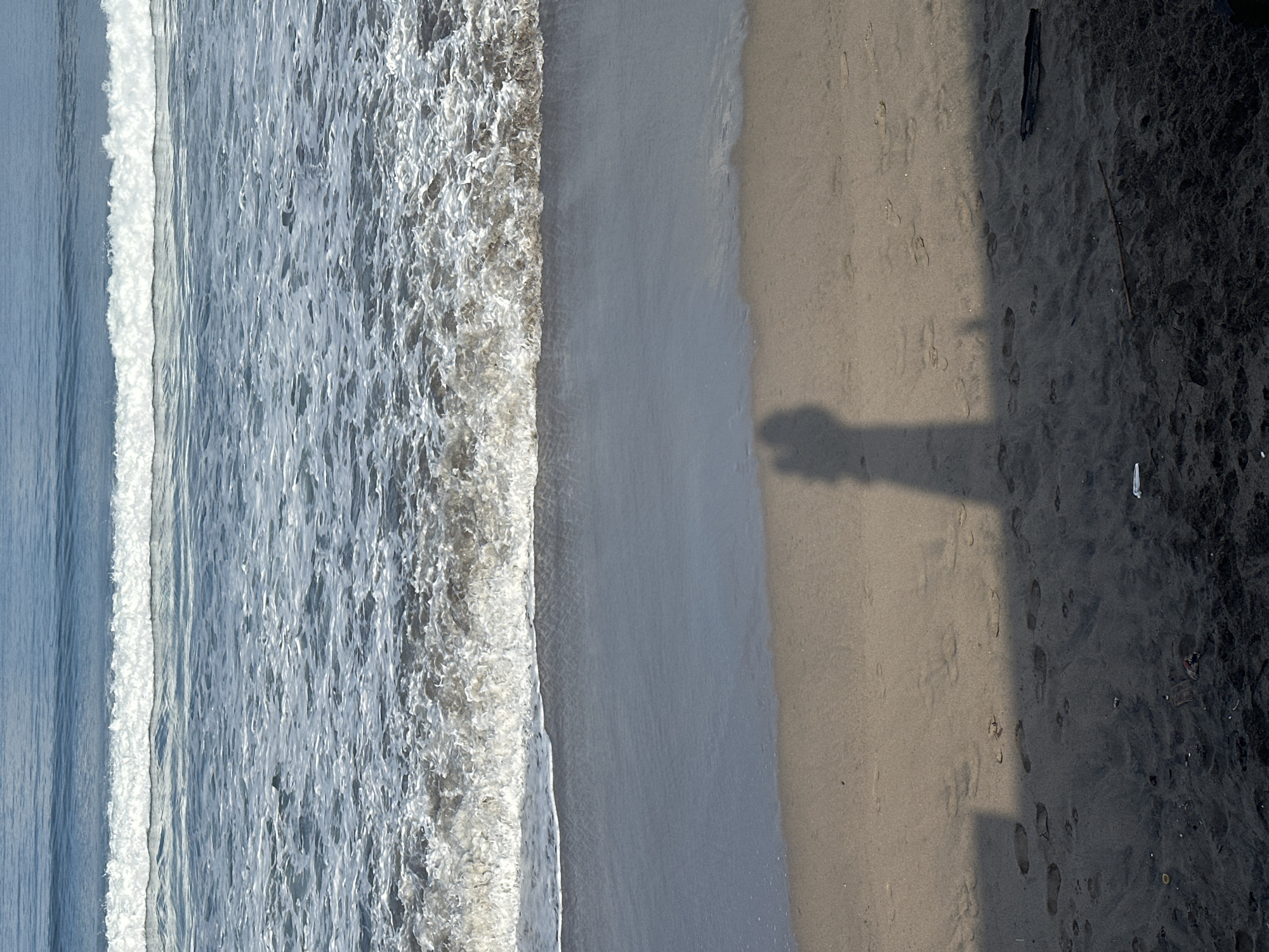 An aerial shot shows the shoreline of a beach in North Kuta, Indonesia. The ocean takes up the left portion of the image, with waves crashing onto the light tan sand. The beach transitions to dark black volcanic sand on the right side of the image. A long shadow stretches across the tan and black sand. There are footprints in both the tan and black sand. A piece of white trash lies on the dark sand. A dark object is situated on the right portion.