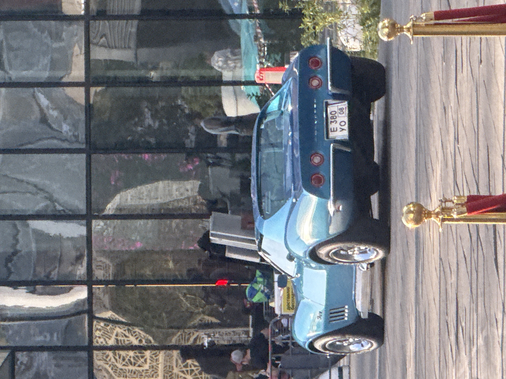 The image captures a blue Chevrolet Corvette Stingray parked on a paved surface, likely a street or plaza, in front of a modern building with a glass facade in Khimki, Russia. The car, viewed from the rear, displays a license plate that reads "E 380 YO 08." In the background, reflections in the glass facade show several people, some silhouetted and others more discernible. One person in the foreground, to the left of the car, is wearing a cap and black shirt. There are also red velvet ropes with gold stanchions in the foreground, suggesting a controlled access area or an event. The environment appears to be urban, with the car and building being the primary focus.