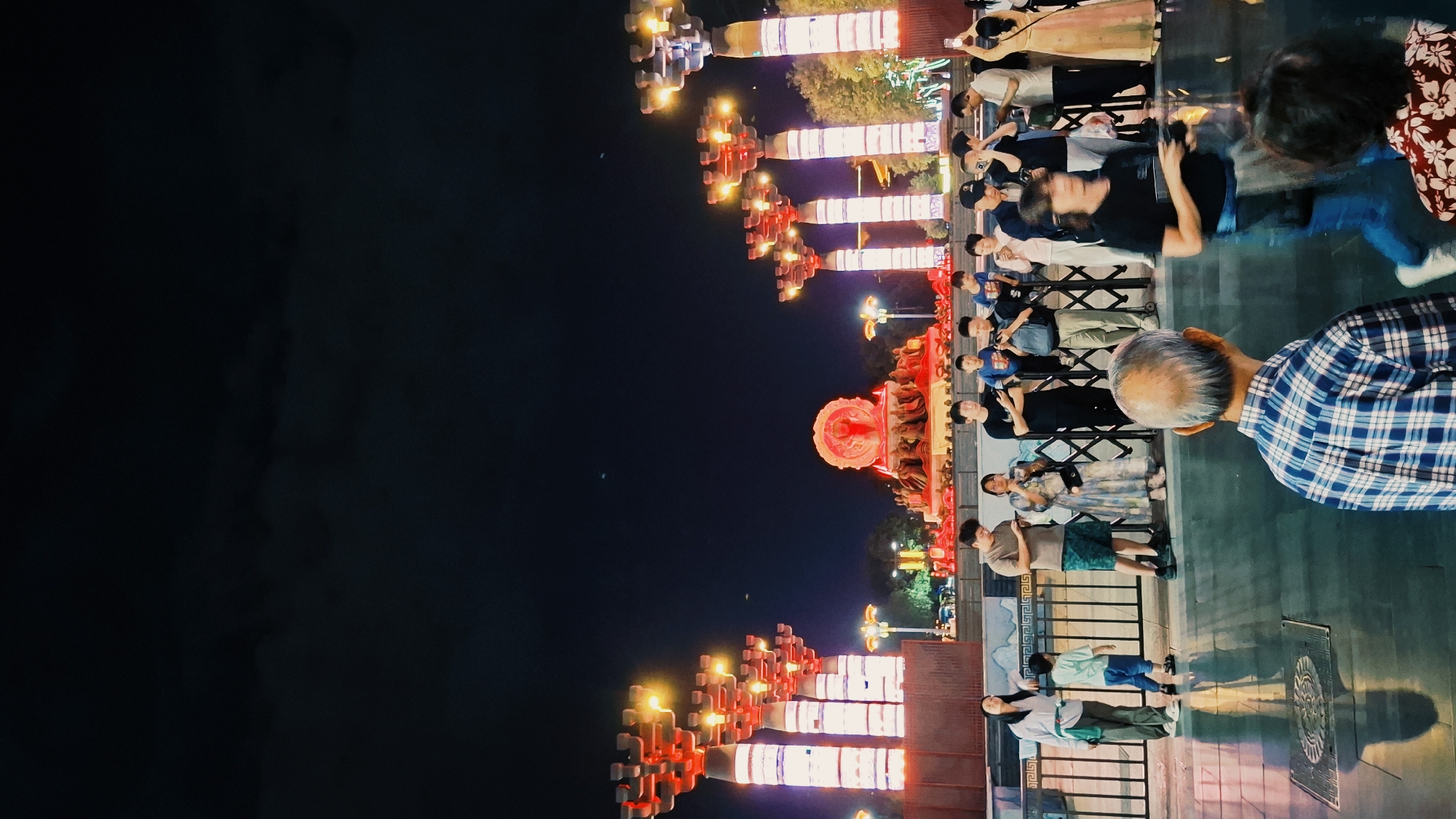 People stand along a railing at night, viewing and photographing illuminated, vertically stacked, red and white architectural displays. The scene is likely located in the Yanta District, China.