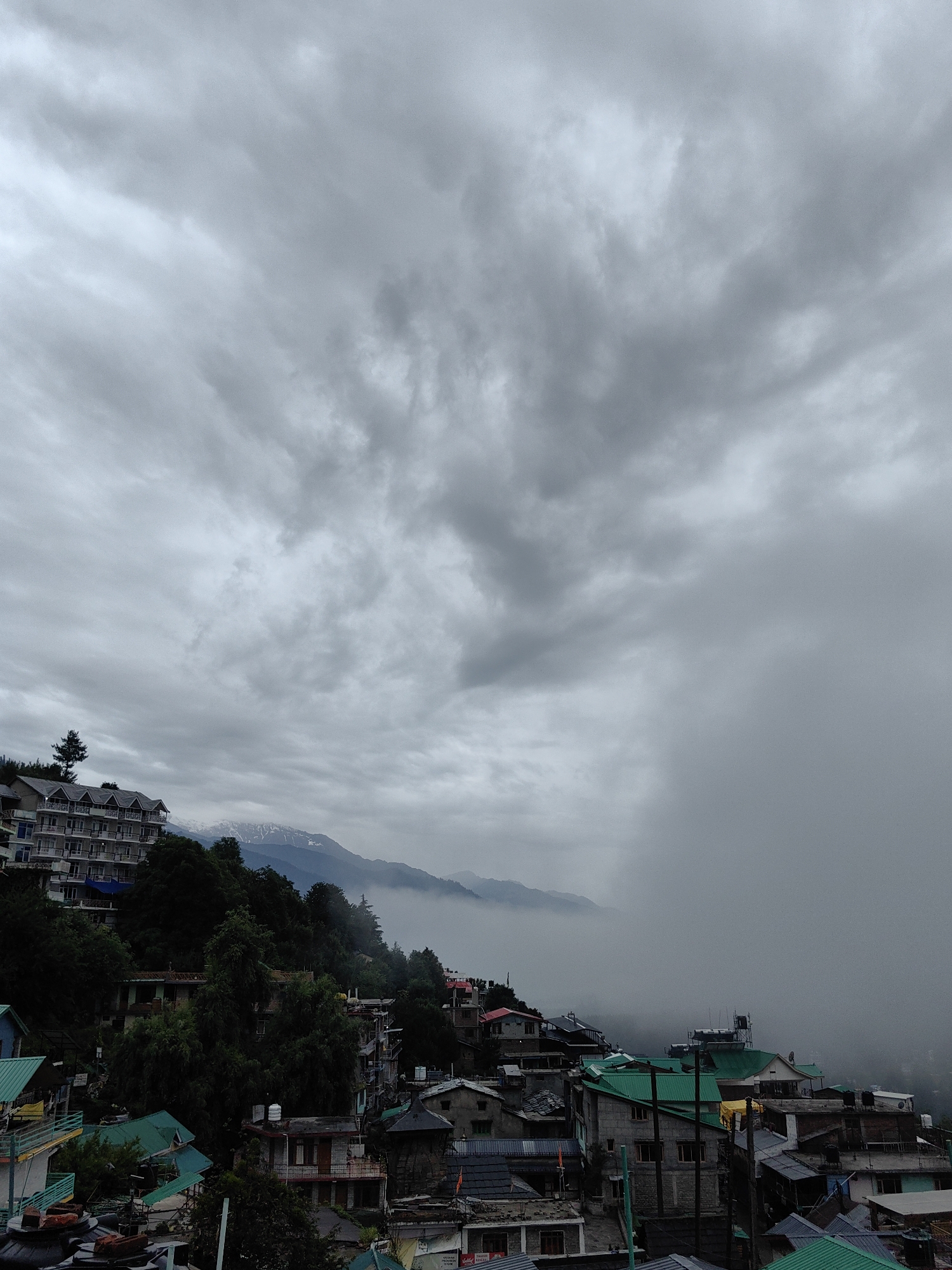 The image depicts an overcast sky over a dense cluster of buildings on a hillside in Vashisht, India. A large hotel or apartment complex with multiple balconies is visible on the upper left portion of the frame, positioned on the slope of a mountain. Further down the hillside, a dense collection of houses and smaller structures with various colored roofs, predominantly green, are tightly packed together. The landscape is characterized by steep inclines and dense foliage. In the distance, snow-capped mountains are partially obscured by atmospheric haze and clouds, which are thick and gray, indicating a cloudy or potentially rainy day. The overall scene conveys a sense of a mountainous village or town under a dramatic sky.
