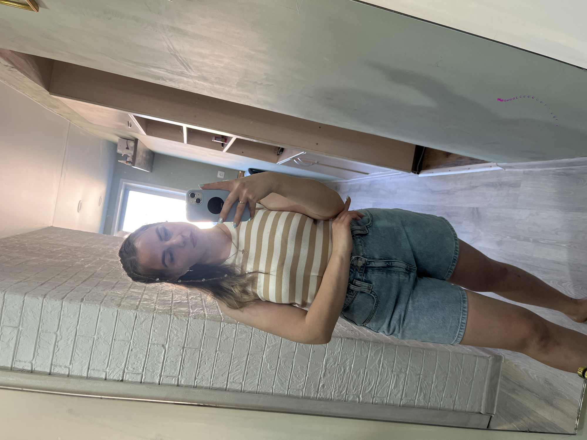 A woman takes a selfie in a mirror. She is wearing a tan and white striped top and denim shorts. The background includes a brick-patterned wall, and a glimpse of a room with shelving and a window. There is no information in the image to indicate this is in Izhevsk, Russia.