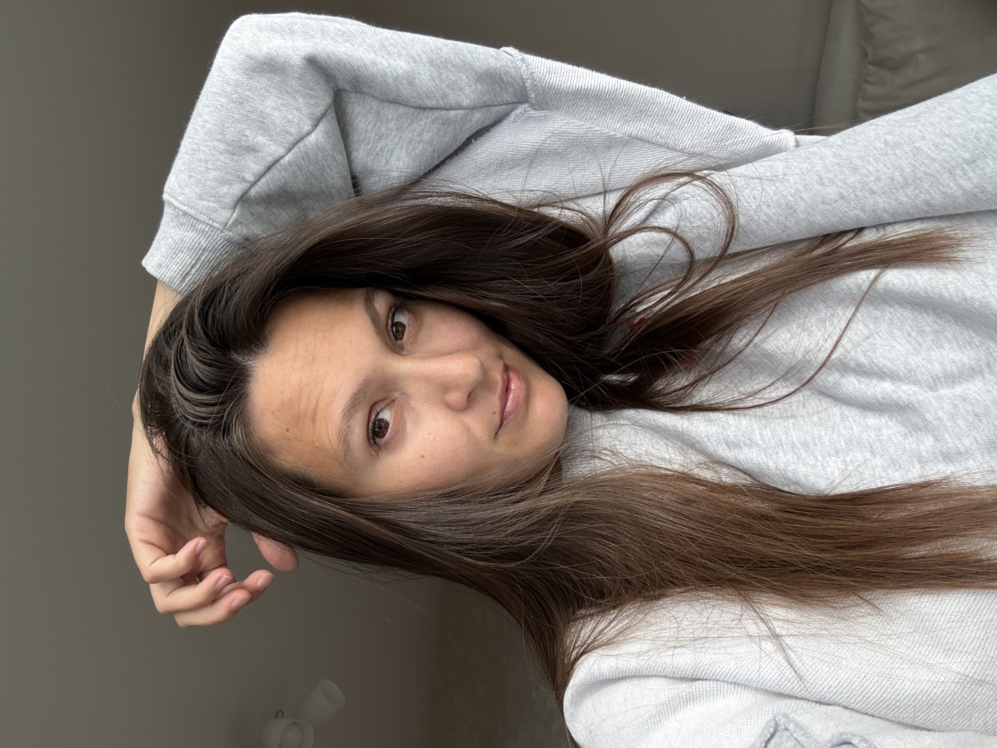 A woman with long, brown hair is posing for a selfie indoors. She is wearing a gray sweatshirt. Her left arm is bent at the elbow with her hand near her forehead. The background includes a plain wall and the edge of a white sofa or bed. It is not possible to confirm the location as the city of Chaikovsky, Russia, based solely on the image.