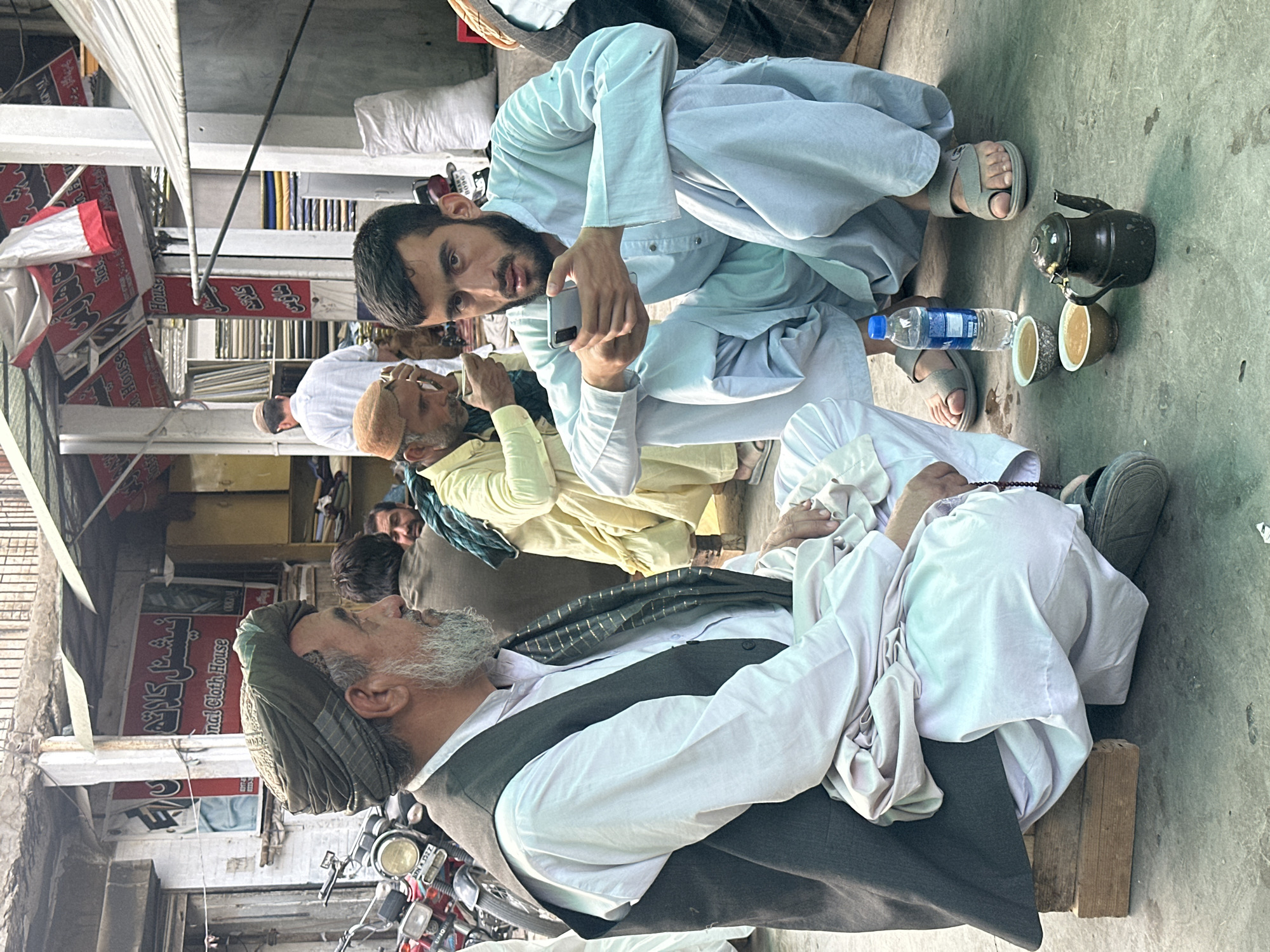 Captured at street level, the photograph presents a tableau of men in Quetta City Tehsil, Pakistan. Predominantly, the subjects are seen seated in traditional Pashtun attire. One individual is actively engaged with a smartphone, while others appear to be in repose or conversation. A tea set, comprising a teapot and cups, is placed on the ground, indicating a shared social moment. The backdrop reveals urban infrastructure, with building facades and commercial signs visible. A motorcycle is also parked nearby, suggesting vehicular access. The image conveys an everyday scene within a populated urban setting.