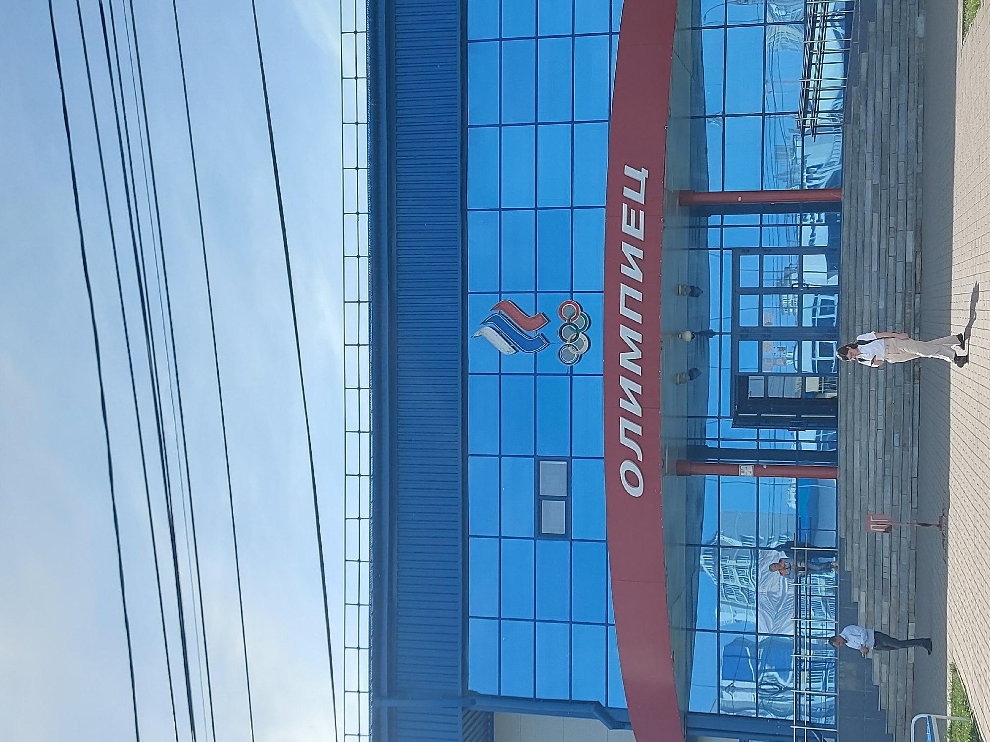 The image depicts the exterior of the "Olympiec" ice stadium in Izhevsk, Russia. The stadium is a multi-story building with a blue facade of square windows. A vertical red banner displays the word "Олимпиец" (Olimpiec) in white letters. The Russian Olympic Committee logo is also visible. Power lines are visible against a bright blue sky in the upper left corner of the frame. Three people are standing on the steps in front of the stadium. The environment appears to be a paved area adjacent to the building.