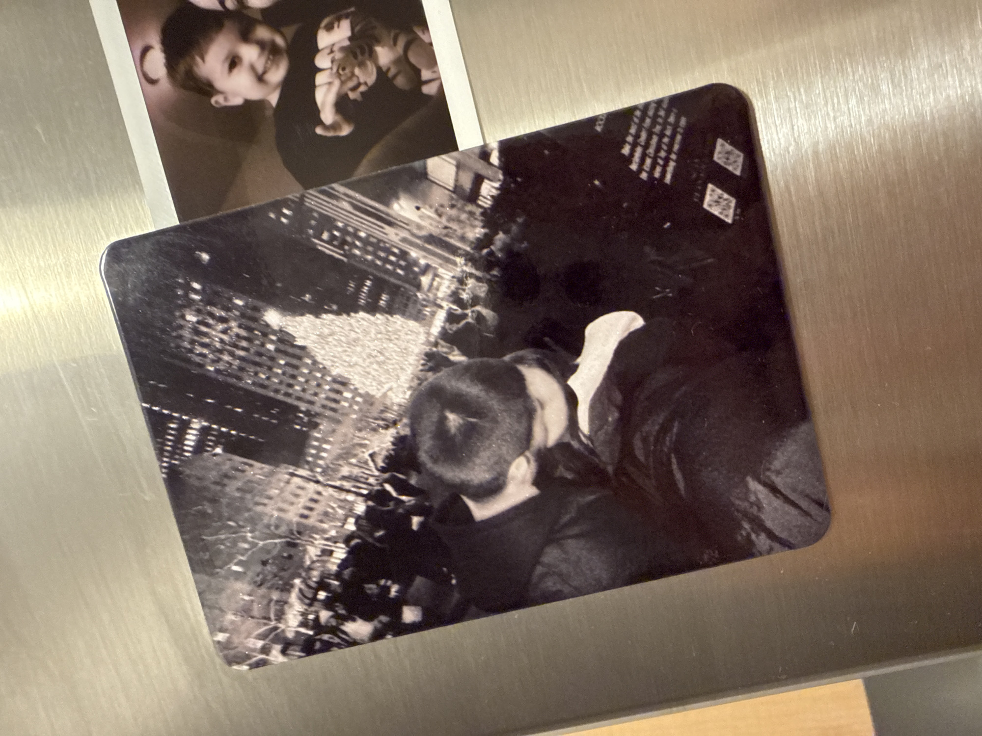 The image displays two photographs on a metallic surface, likely a refrigerator. The larger photograph, on the left, is a black and white image capturing a crowd gathered in front of a brightly lit Christmas tree, with tall buildings in the background. In the foreground of this photograph, a couple is embracing and kissing. The smaller photograph, partially visible on the right, is a color image of a young child smiling and looking towards the camera. The child is wearing a black shirt with a graphic design. The metallic surface shows a brushed texture. The overall scene appears to be an indoor setting, with the photographs mounted on a kitchen appliance. There is no discernible information in the image to indicate the location as the City of Yonkers, United States of America.