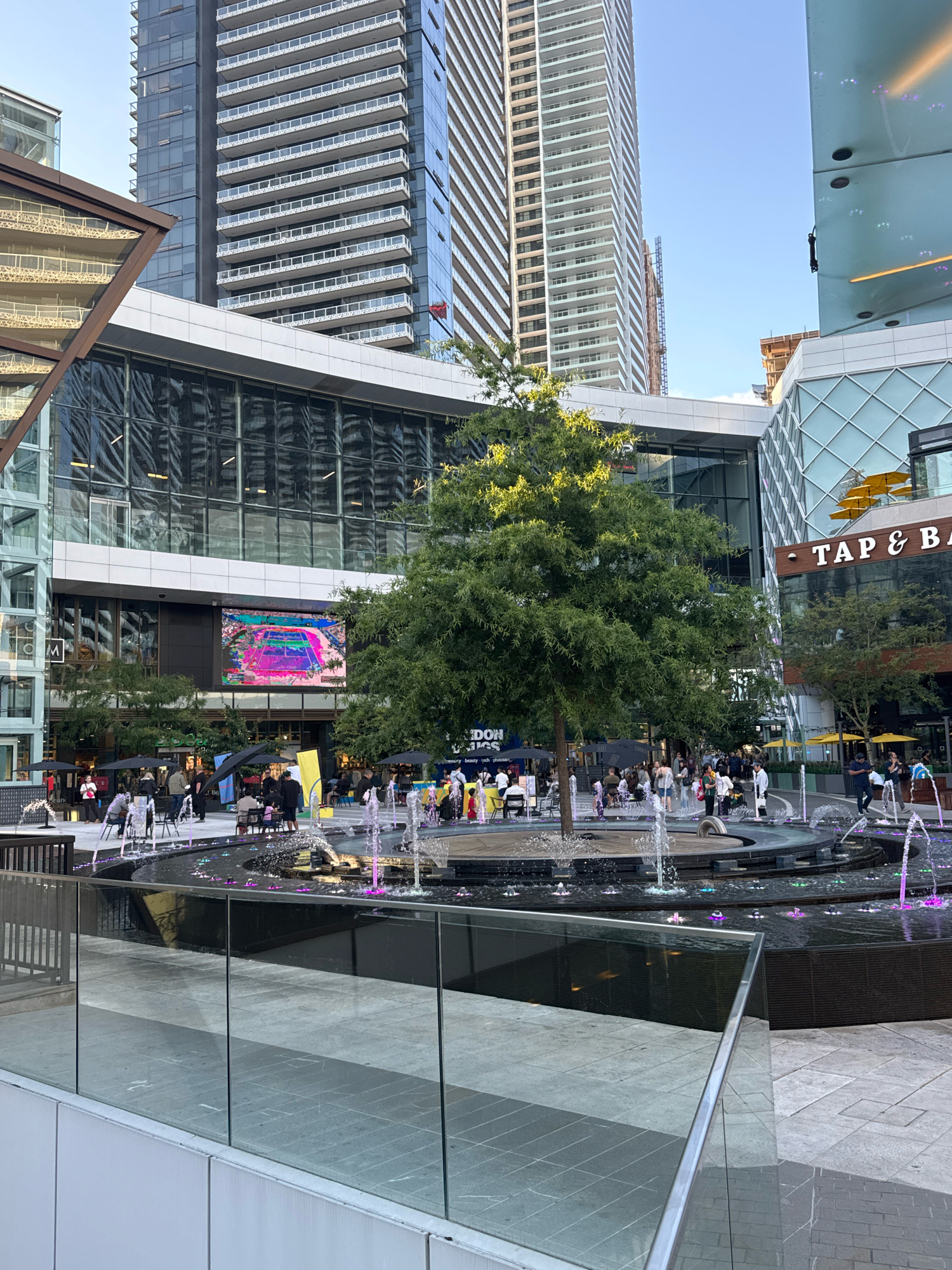 The image depicts a bustling outdoor plaza in Burnaby, Canada, likely at a modern commercial or residential development. The scene is dominated by a large, multi-tiered fountain with several jets of water, some illuminated with colorful purple and pink lights, creating a dynamic and lively atmosphere.
In the background, imposing modern skyscrapers with glass facades and white balconies rise against a clear blue sky, indicative of urban architecture. A large LED screen displays a vibrant, abstract image. Several people are scattered throughout the plaza, some seated at outdoor tables under umbrellas, others standing or walking. The presence of umbrellas and open seating suggests an area with cafes or restaurants.
A prominent tree with lush green foliage stands near the center of the plaza, adding a natural element to the urban landscape. Further background details include storefronts with signage, one reading "TAP & BA," and additional architectural elements of buildings. The overall impression is one of a lively urban public space designed for recreation and social gathering.