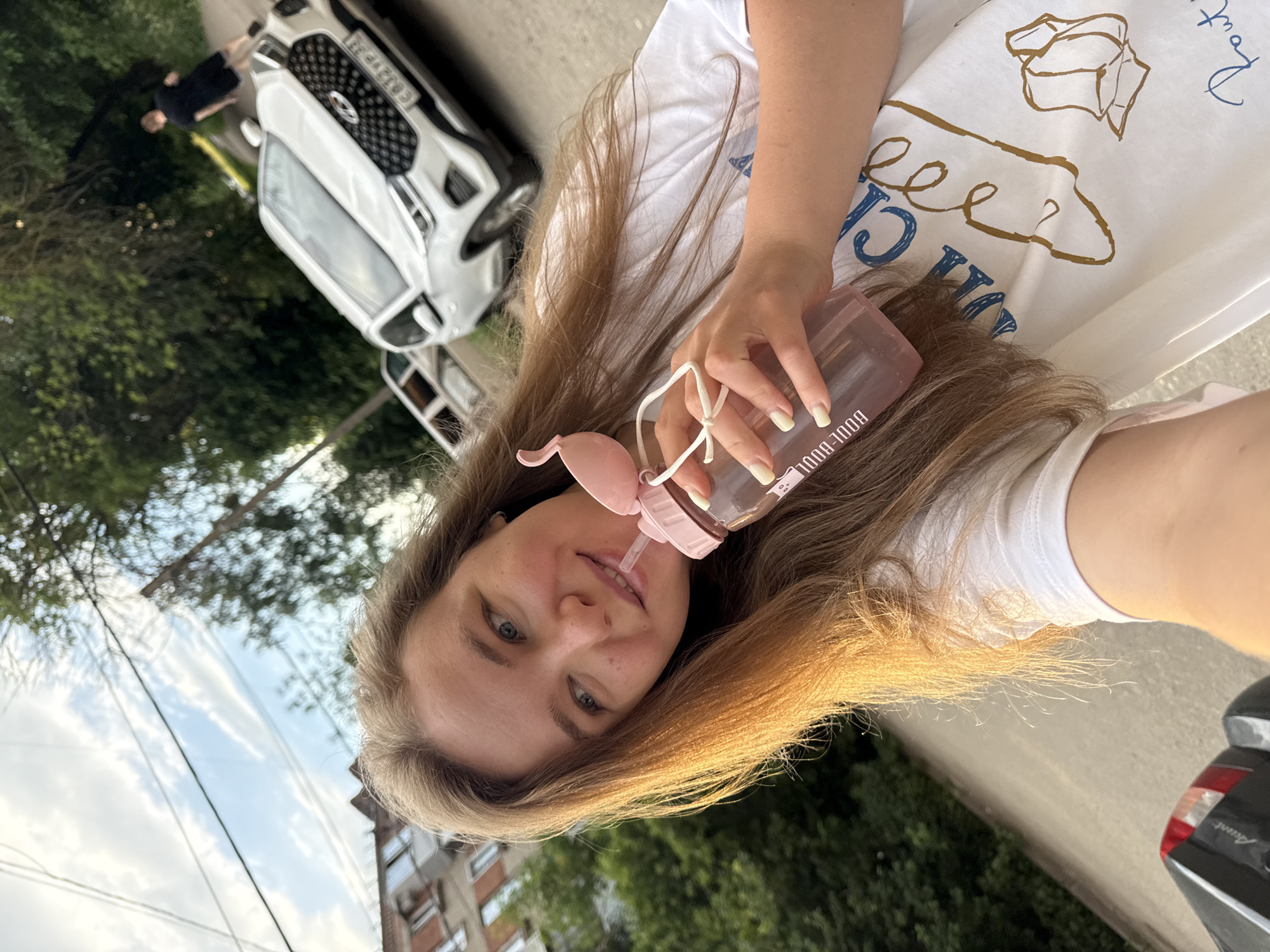 A young woman with long, light brown hair is taking a selfie outdoors. She is wearing a white t-shirt with a drawing on it. She is holding a pink water bottle with a straw to her lips. In the background, there is a white car with a license plate that appears to be Russian. There are trees and a building in the background. The location is the city of Vladimir, Russia.