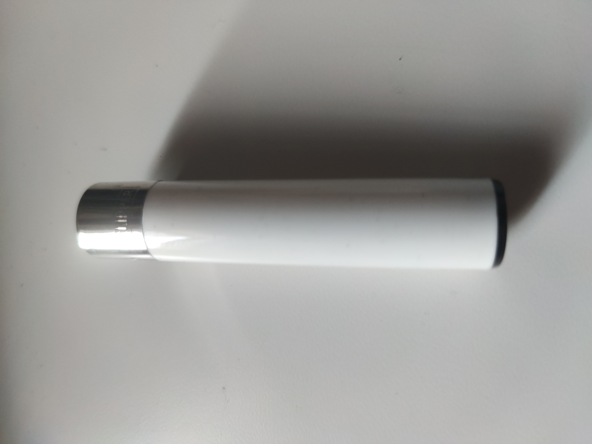 A white Clipper lighter with a silver top rests on a white surface. The lighter is positioned horizontally in the center of the image. The top of the lighter, closest to the left side of the image, is silver, while the body is white. A shadow is cast on the top right of the image. The location is in the city of Unknown, Unknown.