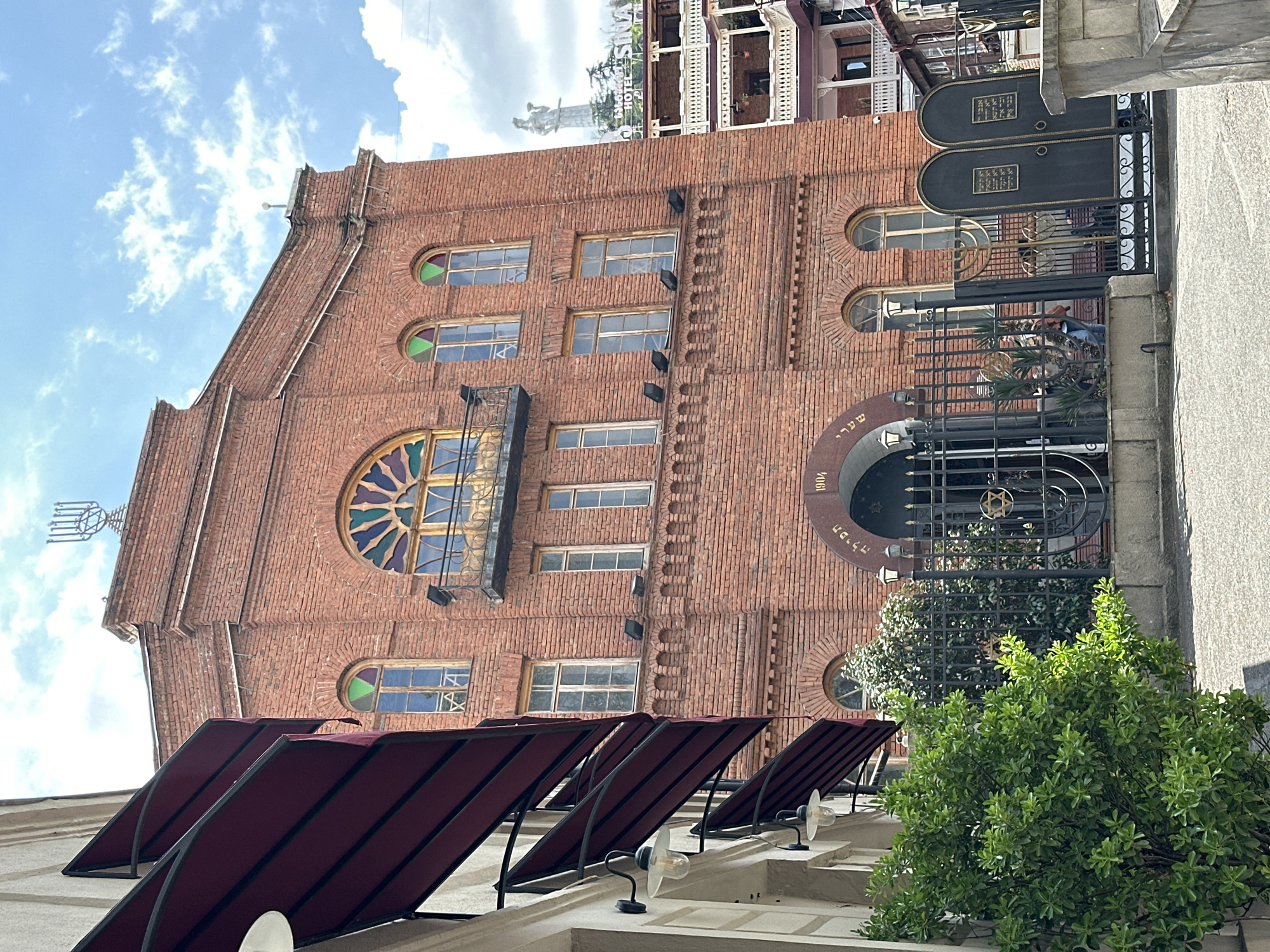 The image captures the facade of a historic brick building, likely a synagogue, under a bright blue sky with scattered white clouds. The building is constructed of reddish-brown brick, featuring varied window designs including rectangular panes, arched windows with colorful stained glass, and a large, ornate stained-glass rosette window above a small balcony.
A prominent metal menorah sculpture is fixed to the left side of the roofline, identifying the building's religious purpose. Above the main arched entrance, Hebrew text reads "שער תפילה" (Sha'ar Tefila - Gate of Prayer) alongside the year "1904." The entrance is guarded by an elaborate black metal gate, which incorporates a Star of David motif and two black panels displaying additional Hebrew script. A person is faintly visible behind the gate.
In the foreground, several maroon awnings extend from an adjacent structure, possibly indicating an outdoor seating area or storefront. Green bushes partially obscure the lower right of the frame. The scene is bathed in bright daylight, suggesting a clear day. In the background, a multi-storied building with balconies and a statue on its rooftop is visible.