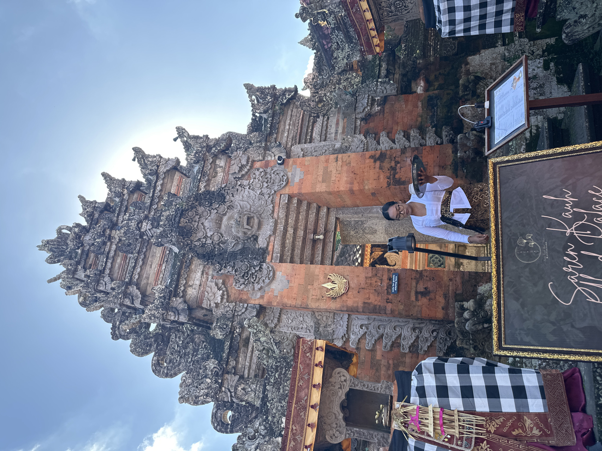 The image depicts the entrance to the Puri Saren Agung Ubud Palace, a historical site in Ubud District, Indonesia. A woman, likely a staff member, stands in front of the ornate stone gateway, holding a silver tray with a small cup. She is wearing traditional Balinese attire, including a white blouse and a patterned sarong. In the foreground, a black framed sign with gold trim displays elegant script, and a menu stand is positioned to the right. The architecture of the palace is intricate, characterized by carved stone deities and colorful fabric draped around the entrance. The sky above is bright blue with some clouds, and the sun is casting a strong light, creating a lens flare. The scene appears to be set on a bright, sunny day, potentially for the arrival of guests or a special event.