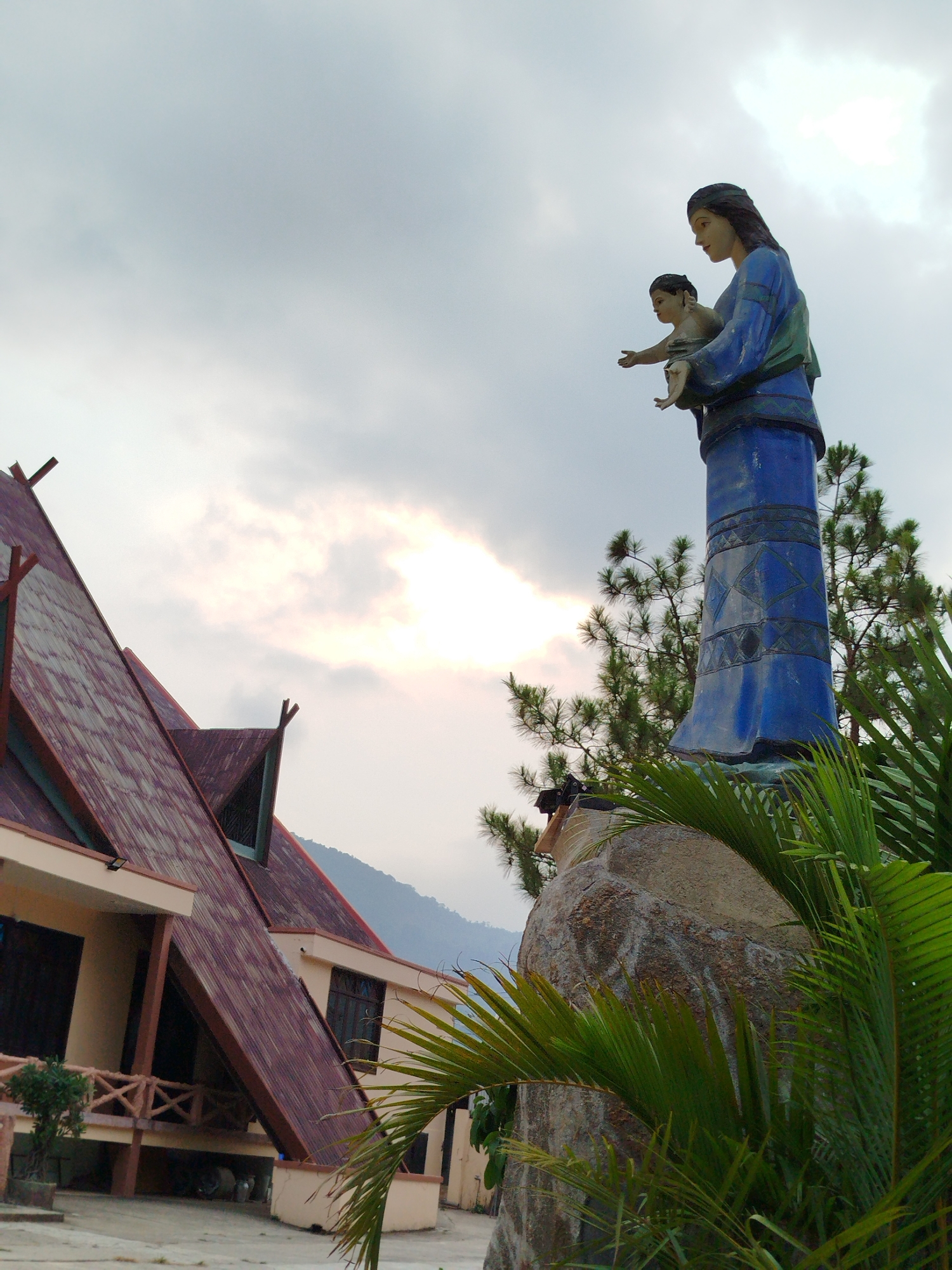 A low-angle shot captures a scene in K'Long, Vietnam, dominated by a statue and a building with unique architecture. The statue, depicting a woman holding a child, stands atop a large rock amidst verdant foliage. The woman is wearing a blue robe, her features distinct. The child is turned outward, his arm extended.
To the left, a building with a steeply pitched roof, reminiscent of traditional Vietnamese architecture, is visible. The roof is a reddish-brown hue. The building has beige walls and windows with dark frames. A small balcony is present, and potted plants decorate the building.
The sky is overcast, with a patch of bright light breaking through the clouds. In the distance, a mountain range is faintly discernible. The foreground features green palm fronds, adding to the tropical feel of the scene.