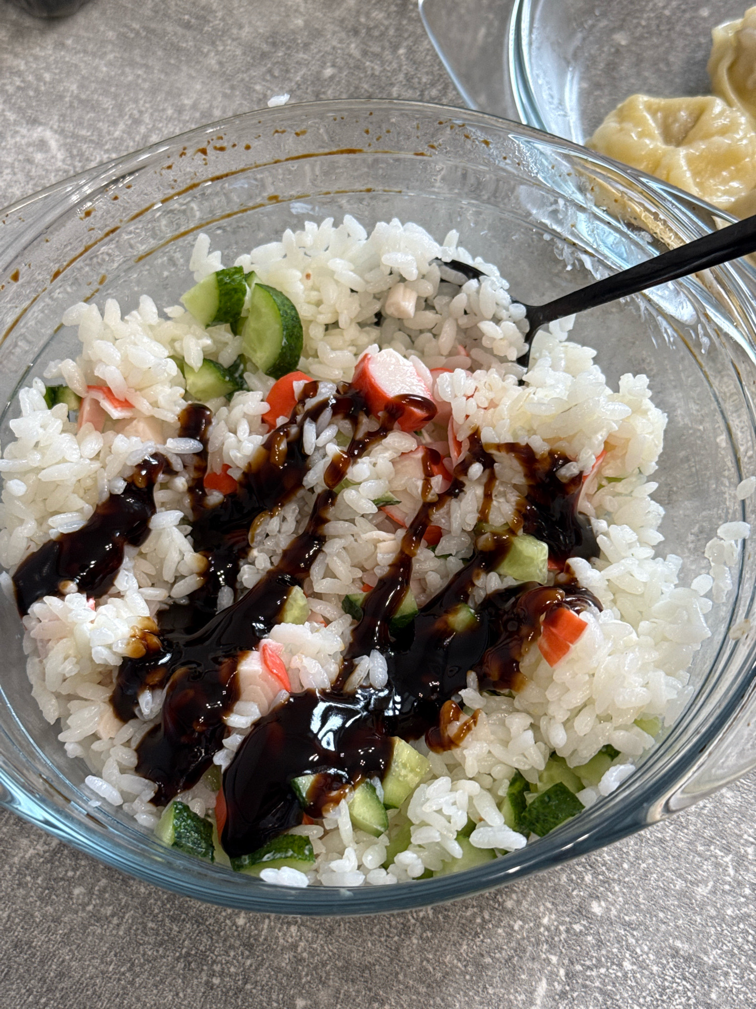 A close-up, top-down shot shows a glass bowl filled with sushi rice, chopped cucumber, and imitation crab meat. Dark brown sauce has been drizzled over the mixture. A black spoon is visible in the bowl. In the upper right corner of the frame, a second glass bowl containing dumplings can be seen. The background consists of a grey speckled surface, likely a countertop. The scene is indoors and appears to be a kitchen environment, with no specific landmarks in Lyubertsy, Russia, identifiable.
