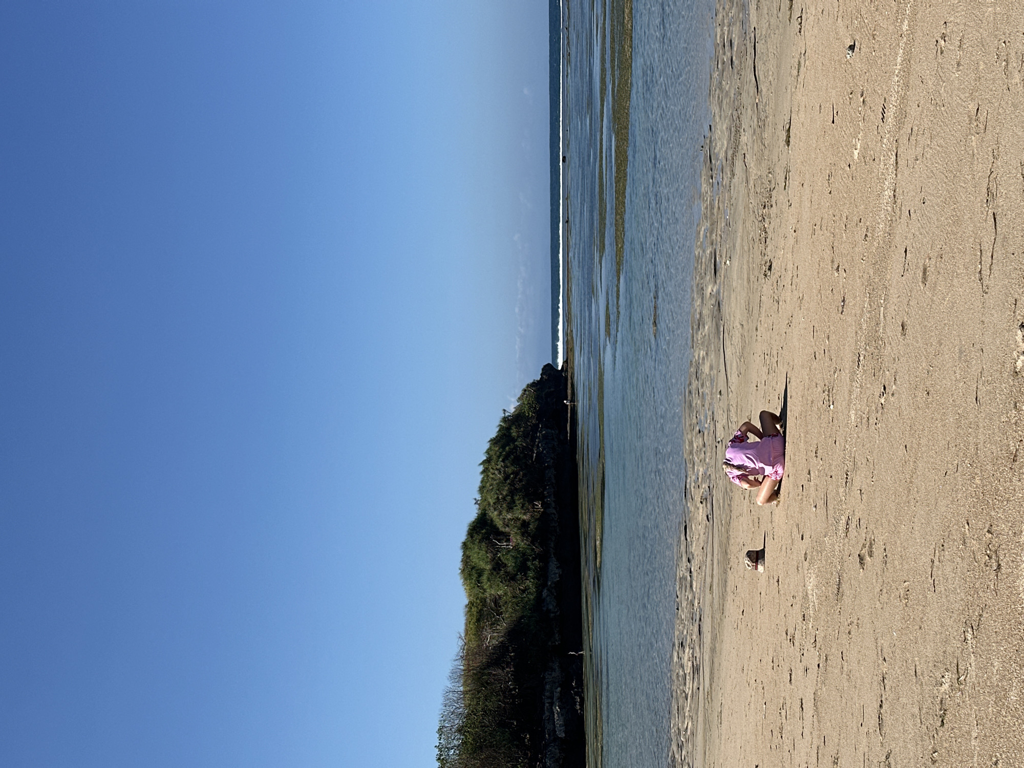 A person wearing a pink outfit sits on a sandy beach in Kuta Selatan, Indonesia. The person is facing away from the camera, with their head bowed. A hat lies on the sand next to them. The beach has wet sand near the water, with receding tide pools visible. In the background, there is a body of water with a gentle wave breaking in the distance. A vegetated cliff face forms the horizon to the left. The sky is clear and blue.