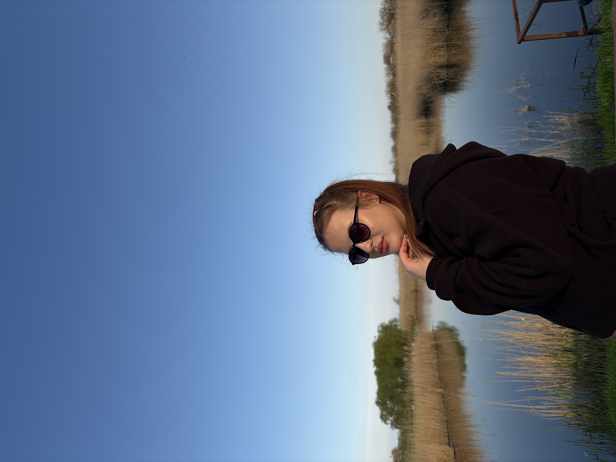 The image shows a young woman posing outdoors against a serene natural backdrop, likely in Novopokrovskaya, Russia. The woman, visible on the right side of the frame, has light brown hair pulled back with a pink hair tie. She is wearing dark sunglasses and a black hooded fleece jacket, resting her right hand thoughtfully on her chin. Her gaze is directed slightly to her right.
The setting features a calm body of water in the middle ground, which reflects the clear blue sky above and the surrounding vegetation. The far bank is dominated by extensive patches of dry, golden reeds or tall grasses, suggesting a late season such as autumn or early spring. A lone, verdant tree stands prominently on the distant shoreline. Closer to the foreground, lush green grass lines the water's edge. A partial view of a rustic wooden structure, possibly part of a dock or support, is visible on the far right of the frame, extending into the water. The sky is a vast, unblemished blue, indicating clear, sunny weather, likely late morning or early afternoon. The overall mood is tranquil. No text is visible in the image.