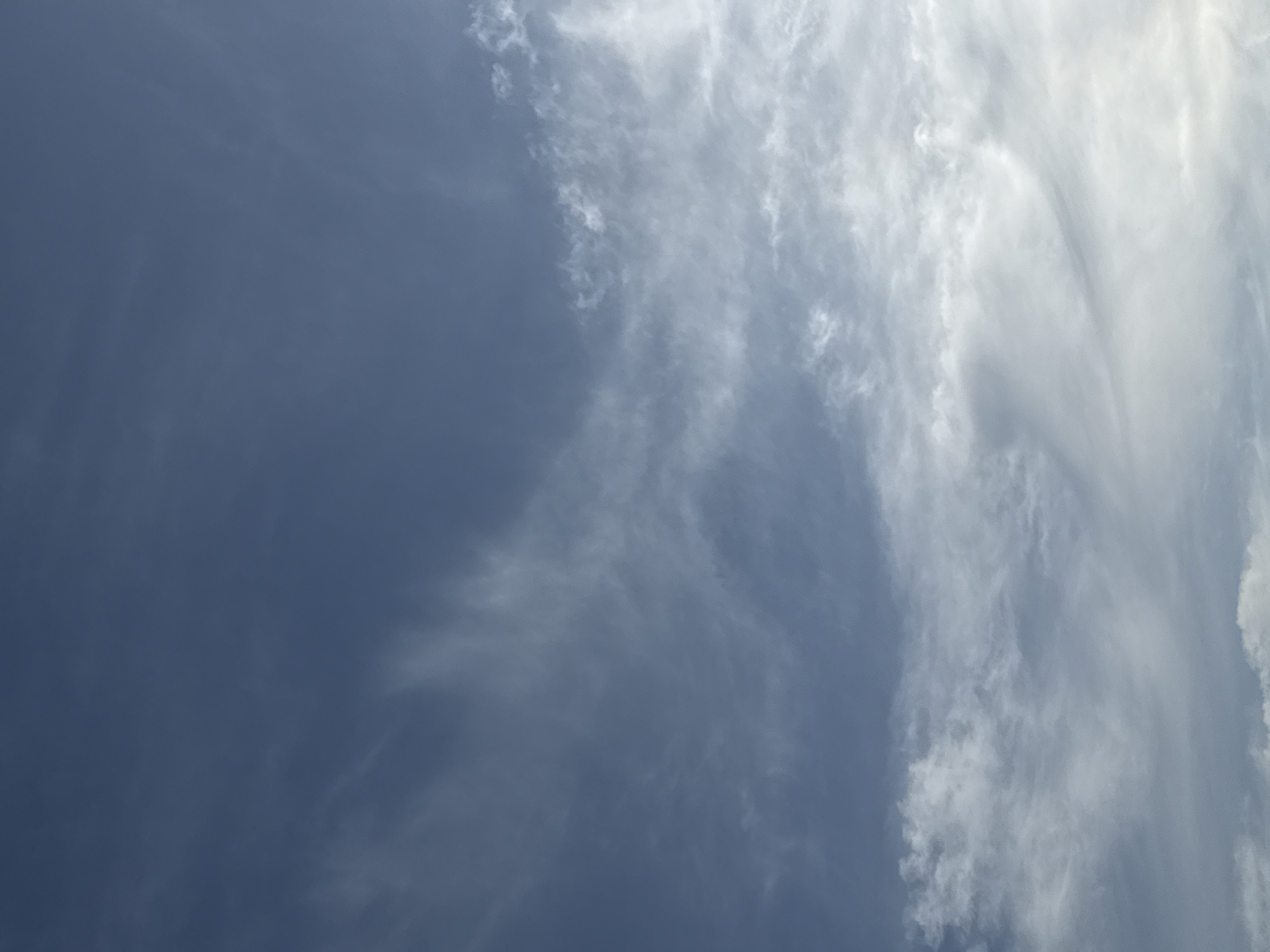 The image depicts a sky with varying cloud formations. The upper portion of the image is a deep blue, indicating a clear sky, while the lower half is filled with wispy, white clouds. Some of the clouds are thin and feathery, appearing to be cirrus clouds, while others are denser and more scattered. The lighting suggests it might be daytime, possibly late afternoon or early morning, given the softness of the light and the coloration of the sky. There are no discernible landmarks or structures visible, making it impossible to determine a specific location within a city or any urban context. The scene is entirely natural, focusing solely on the atmospheric conditions of the sky.